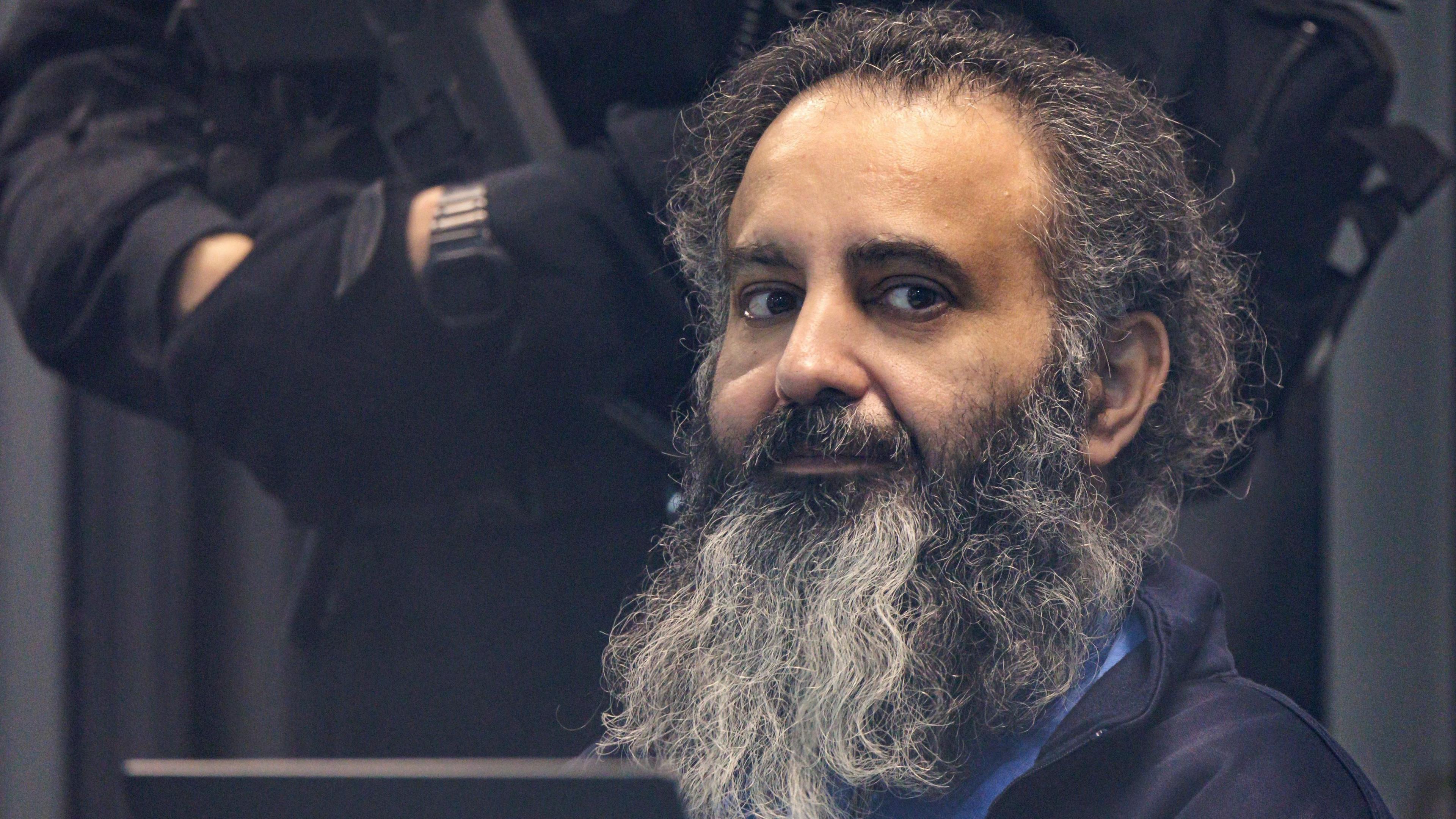 Taleb A. He has a big black and grey beard and fairly long dark hair, streaked with grey. Behind him parts of an armed police officer dressed in black can be seen