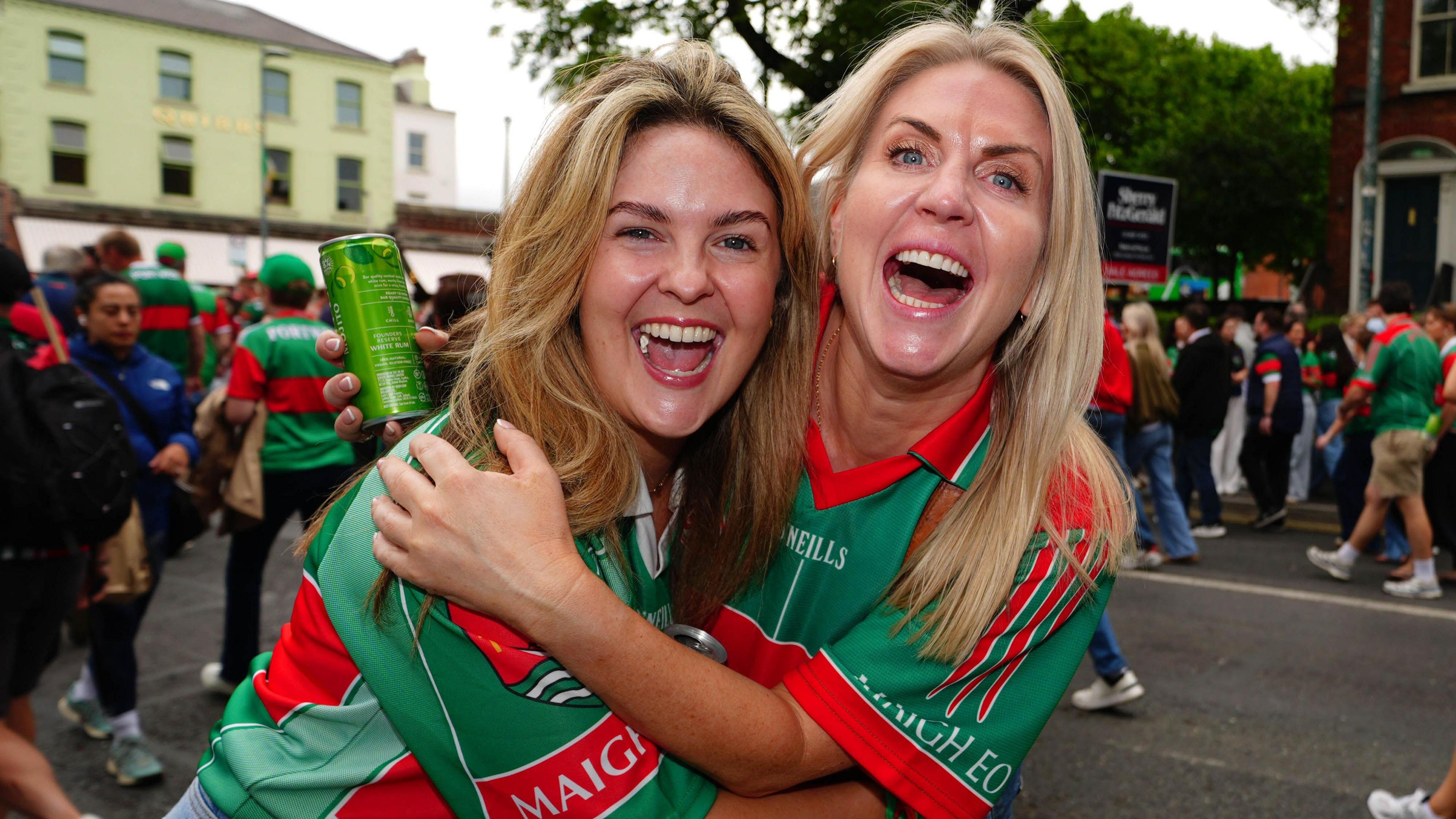 Two people stand in the street, excitement on their faces. They both have blonde hair and are wearing green and red jerseys. Behind them there is a crowd of people walking, many of whom are also wearing green and red jerseys. 