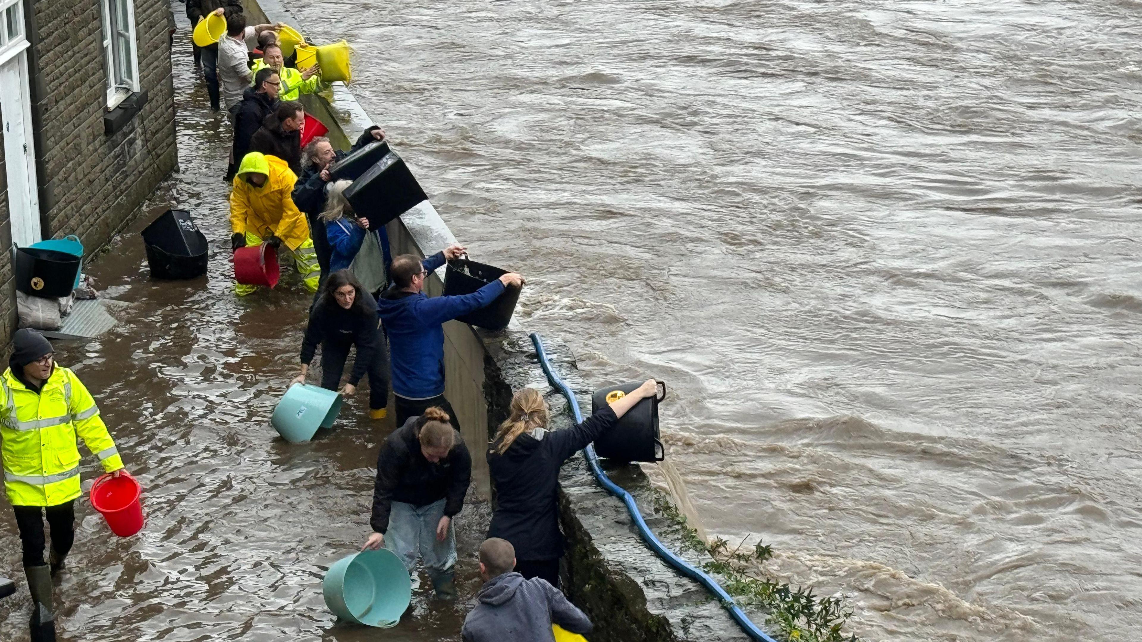 A group of people stand beside a swollen river on a partially submerged path and use buckets to bail water 