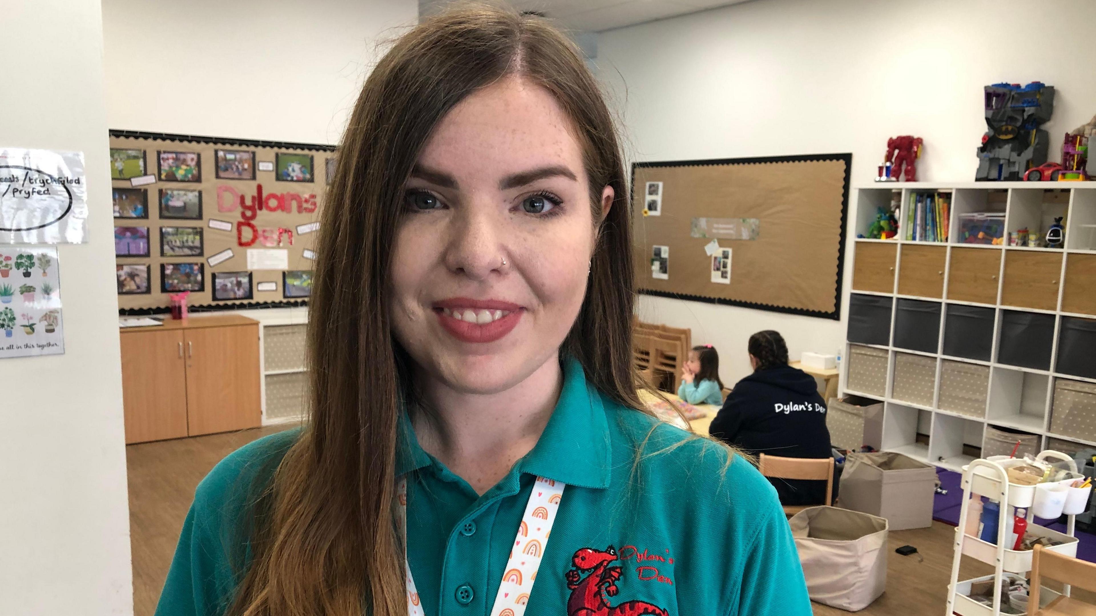 Sarah White wearing a blue polo neck with a red Dylan's Den logo which includes a dragon and a white lanyard with rainbows on it. She is standing in a play room with display boards and a colleague is sat in the background with a child