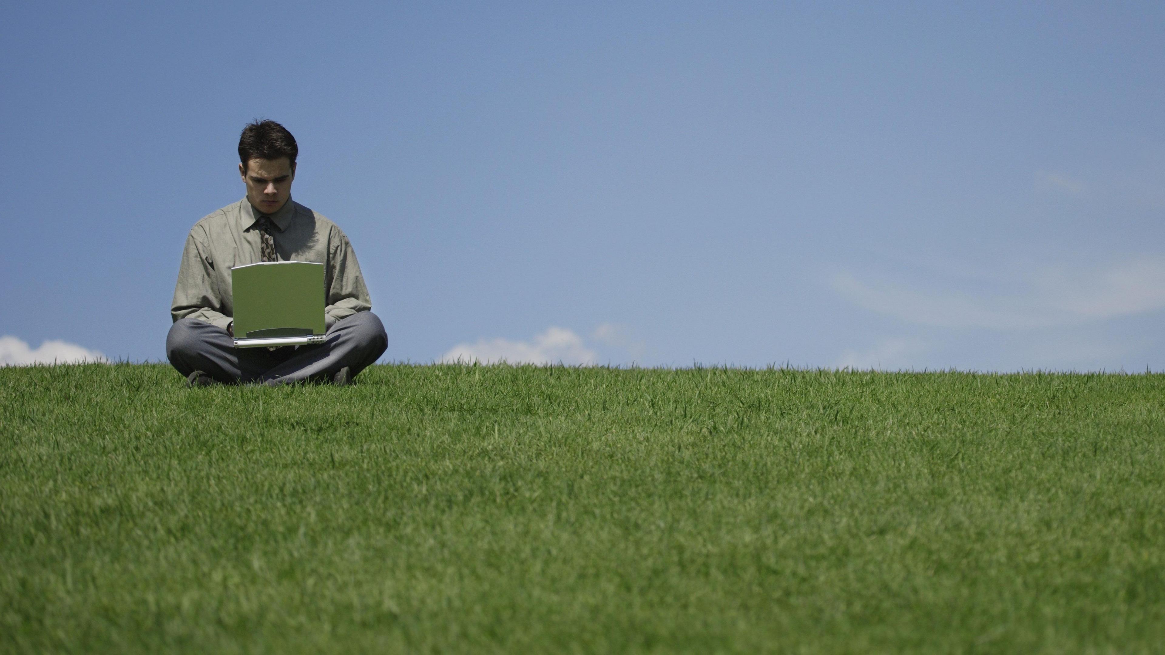 A young man is sitting cross-legged on the brow of a grassy hill typing on a laptop. He is wearing trousers, a shirt and a tie and his head is bowed as he types. It is a clear blue sky with a couple of wispy clouds.
