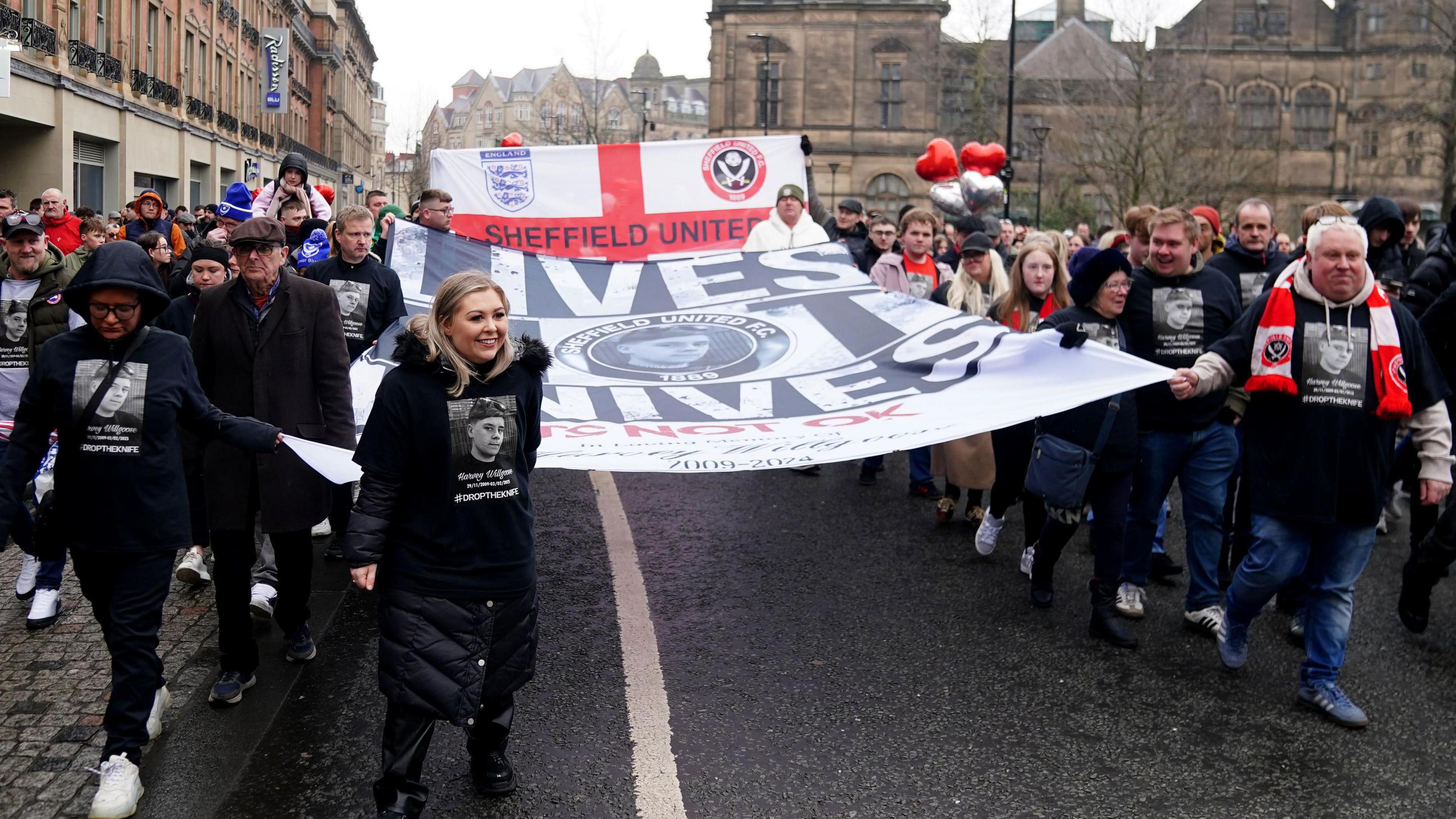 A crowd of people wearing black tops with a picture of a boy on them. They are walking down a street carrying large banners - one says 'lives not knives' and the other is an England flag that says 'Sheffield United'