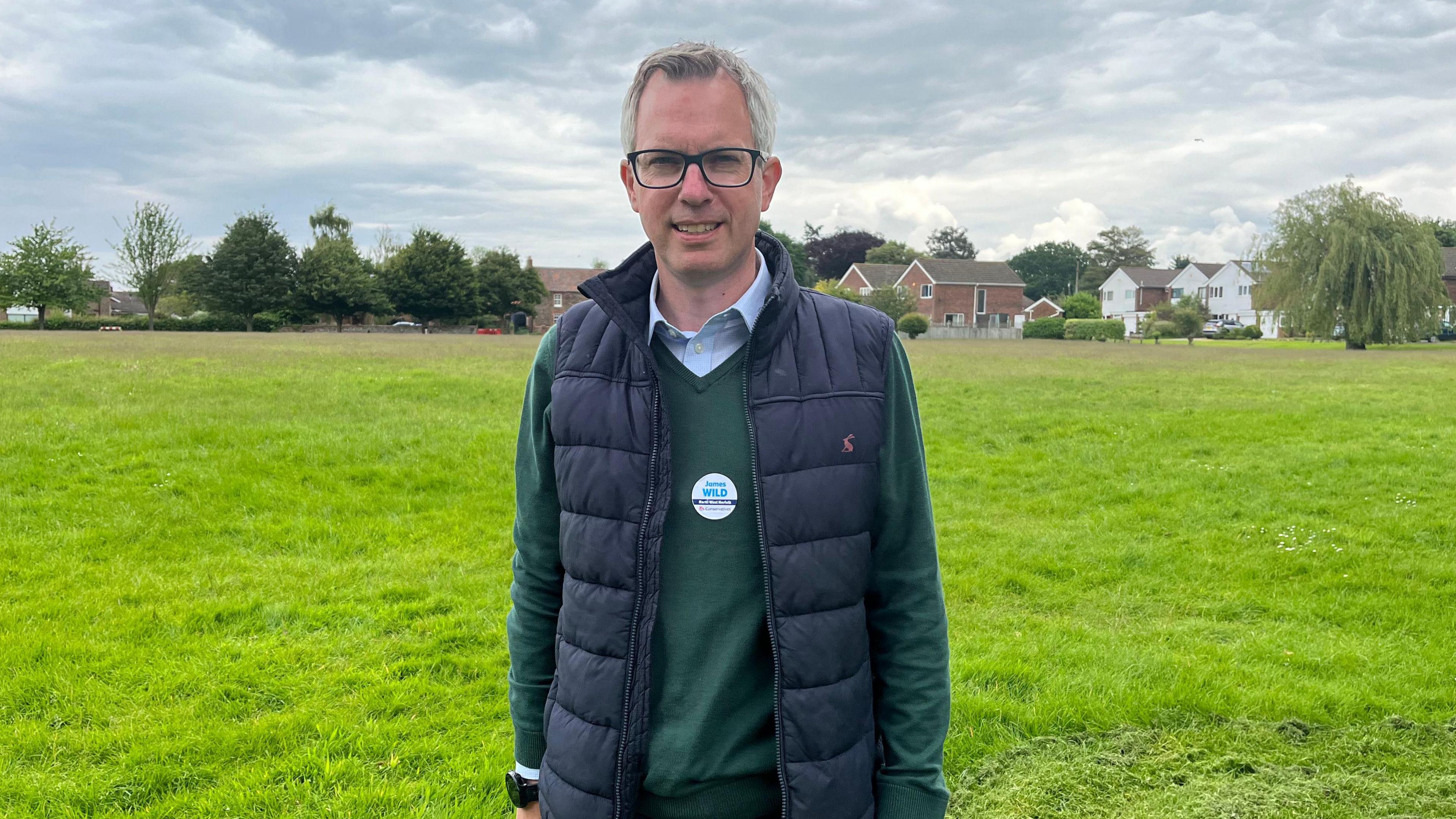 Conservative prospective parliamentary candidate for North West Norfolk James Wild stands on a village green wearing a "vote conservative" sticker and a padded gilet over a green sweater