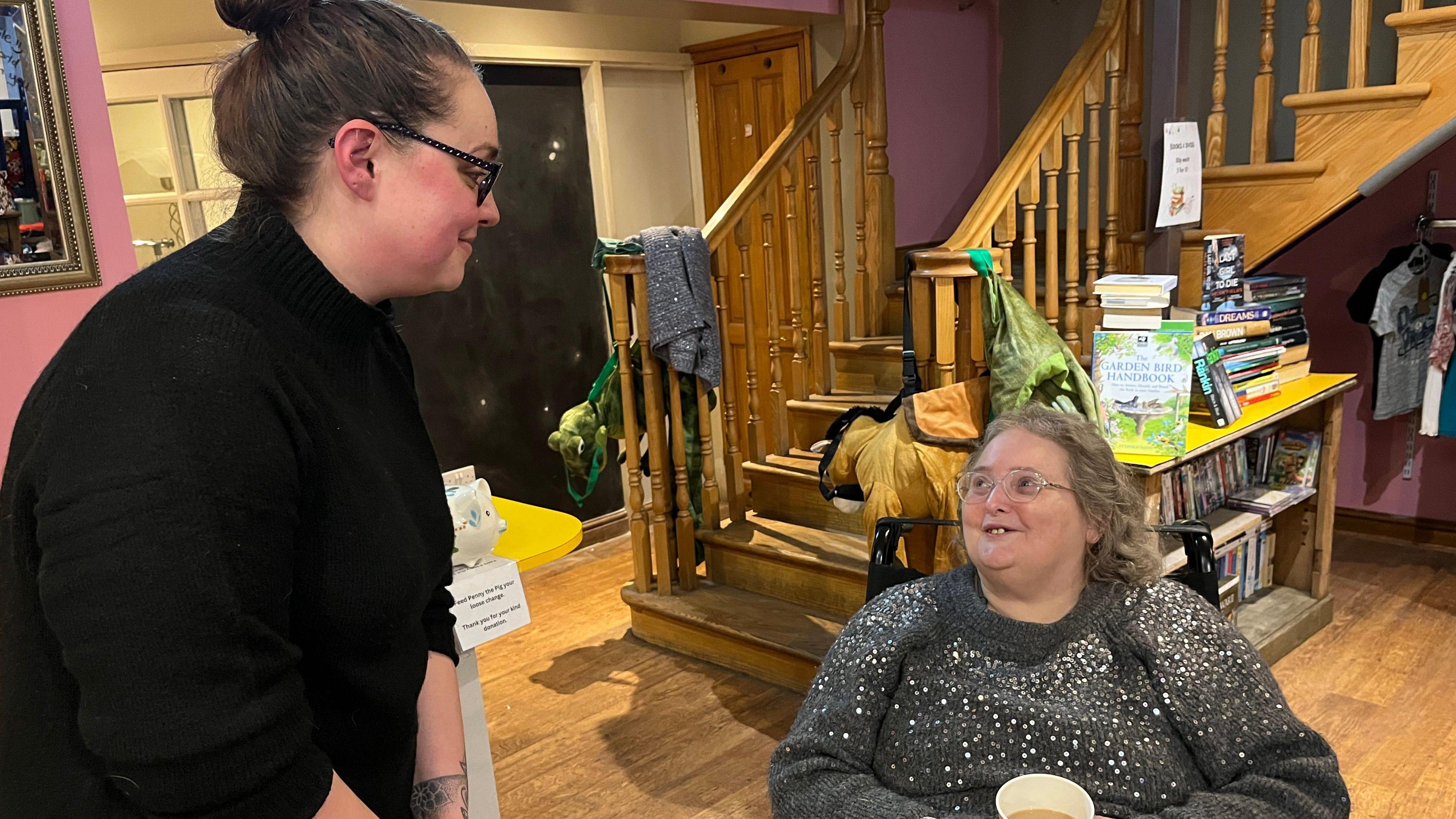 Lisa and Paula Myatt in the Communitea hub in Wigton. Lisa is standing next to Paula, who is in a wheelchair. Lisa is wearing a black jumper, while Paula wears a silver and sequin jumper. Both wear glasses. Paula is holding a cup of tea. 