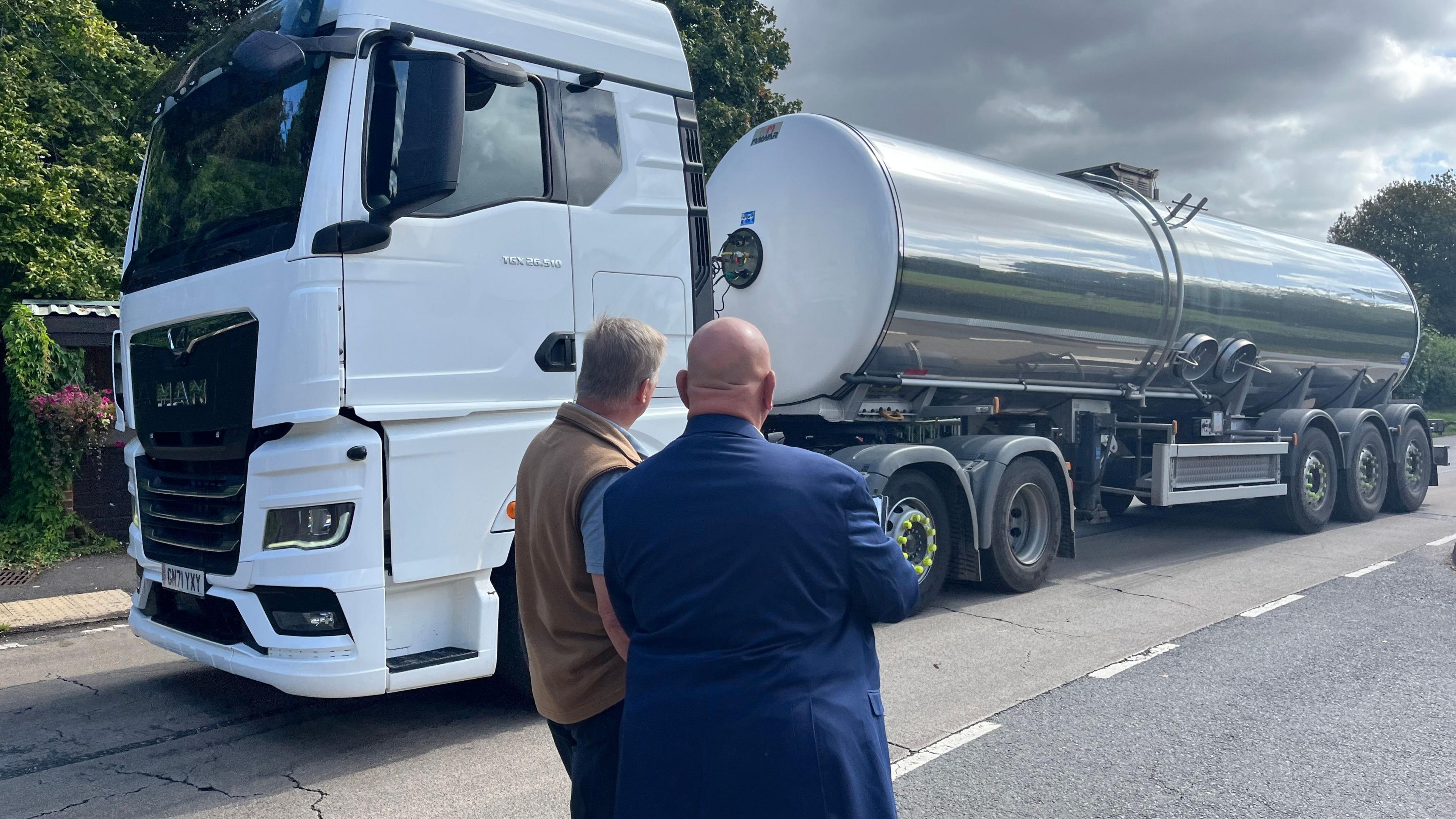 A water tanker drives past two men who watch by the side of the road. 