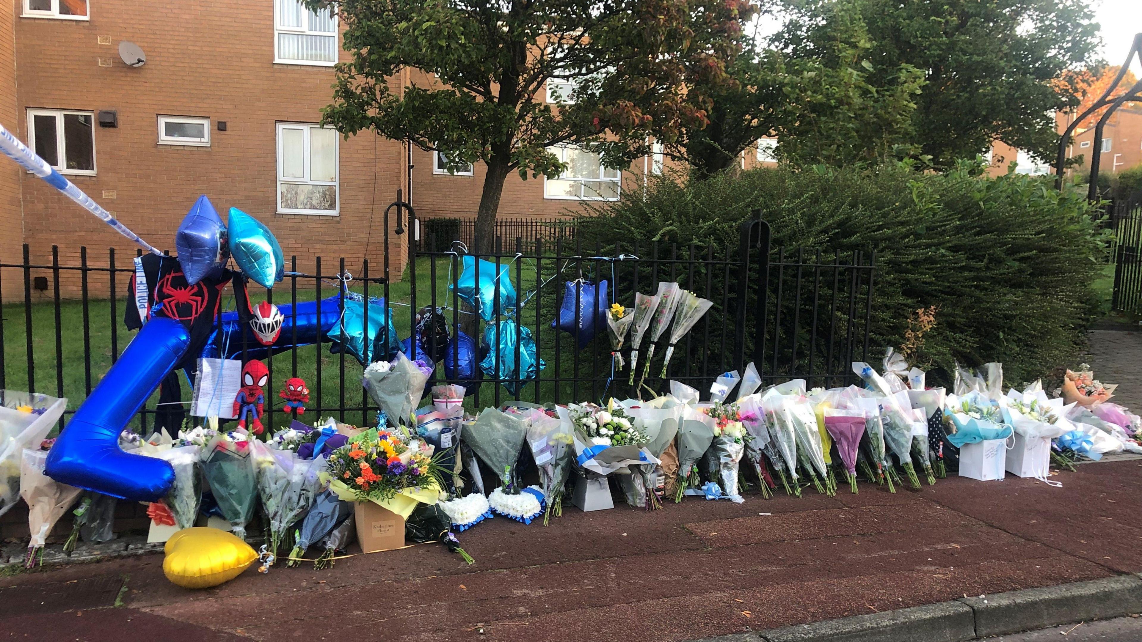 Dozens of flowers and balloons have been placed next to a black fence which surrounds a housing estate. To the left, the police cordon can be seen.