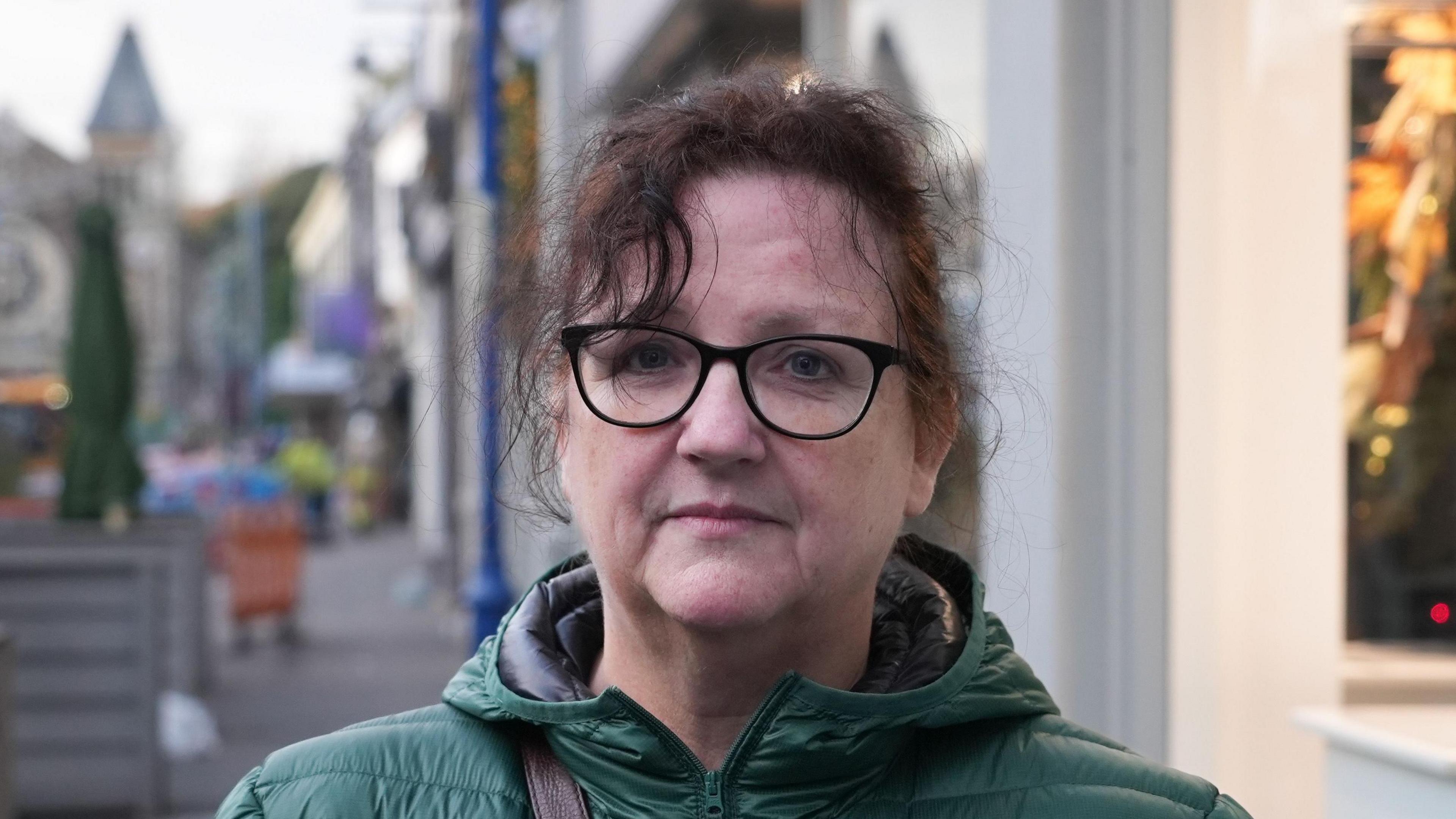 Lesley Reynolds standing in a town street. She is wearing dark glasses and a green puffa coat. Her dark hair is tied up. 