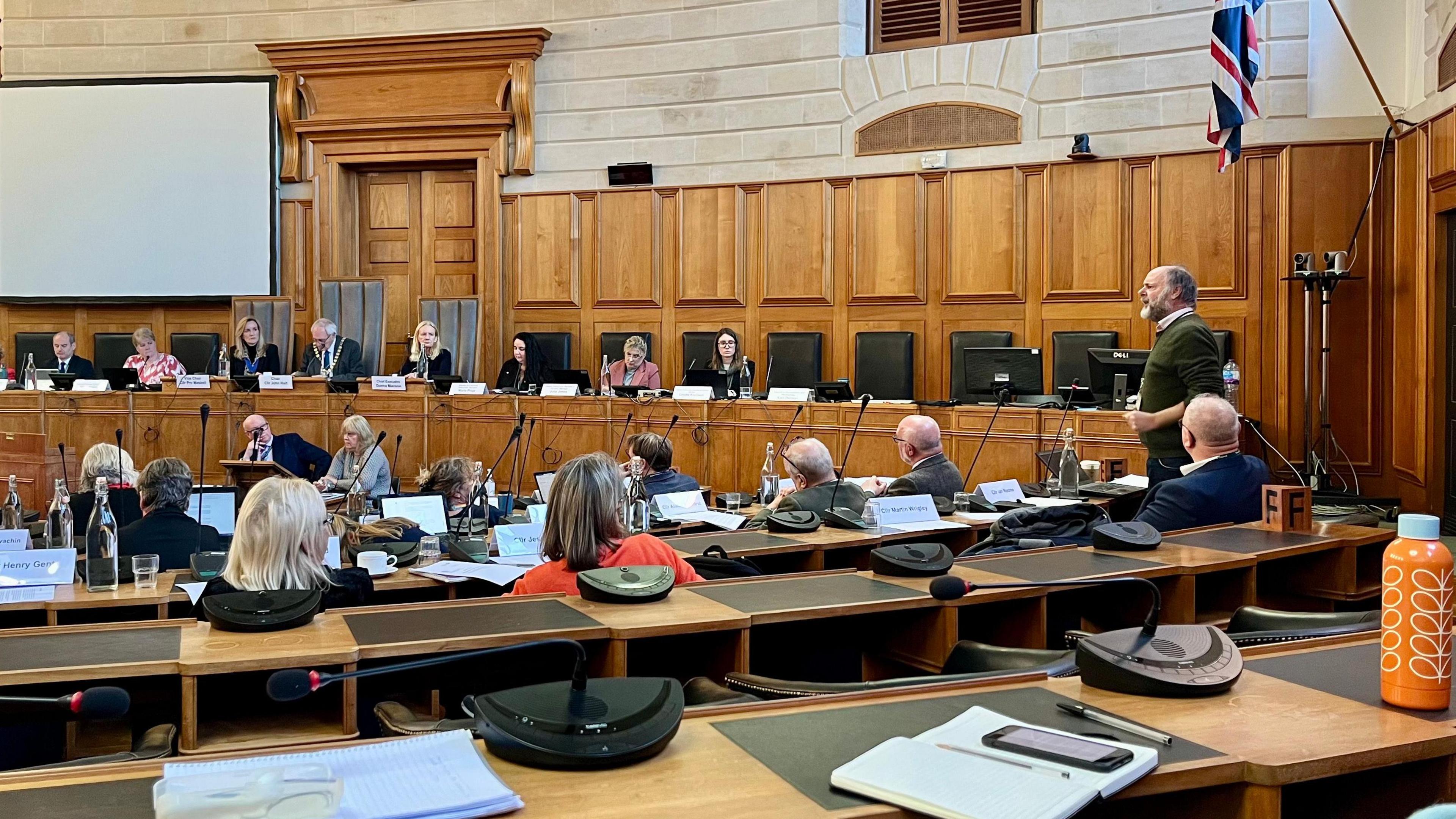 Julian Brazil standing and addressing the chamber at County Hall. Councillors are seated behind microphones in a wood panelled room. The union flag hangs from one of the walls