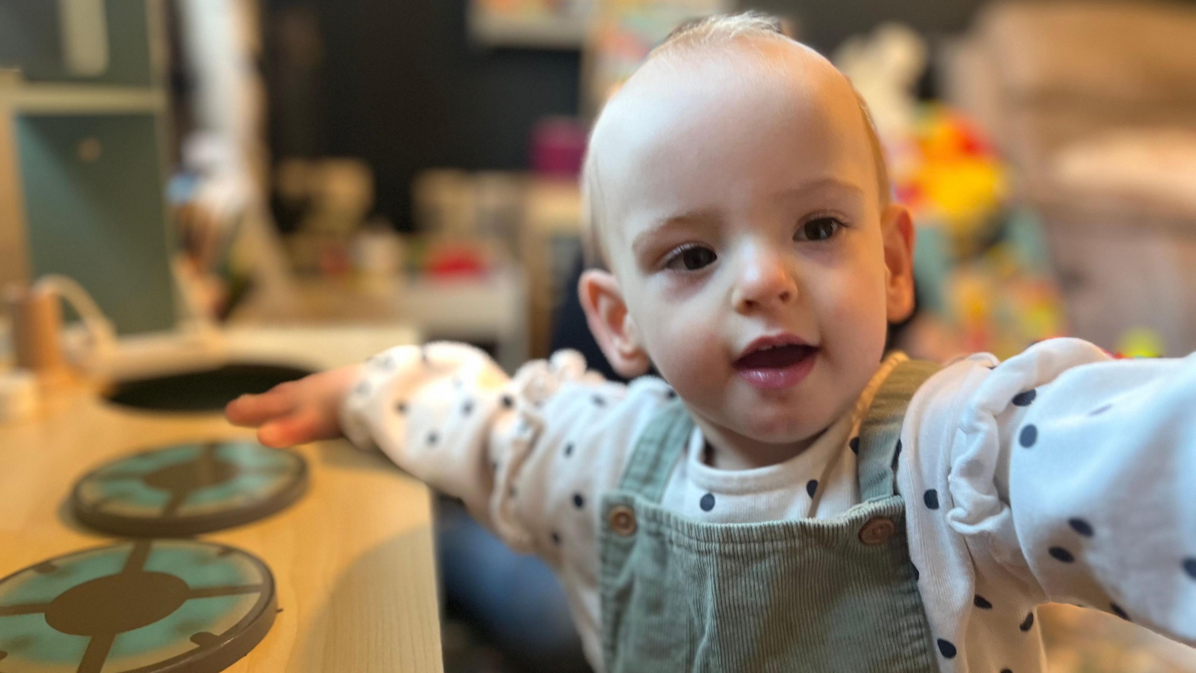 Wren standing up against a wooden table, wearing a green pinafore and white shirt with black dots