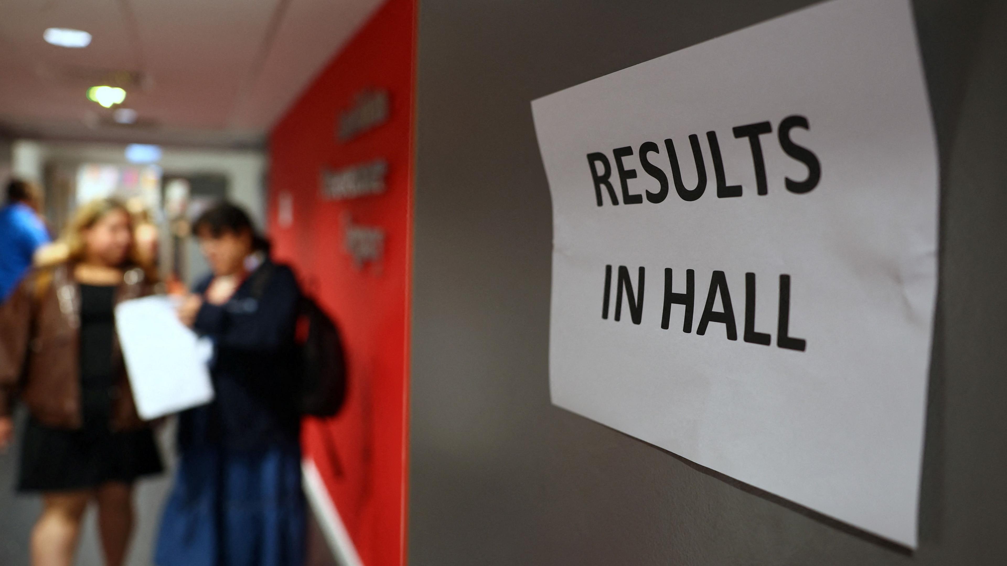 Students, blurred out, stand in the background of a sign that reads "Results in hall"