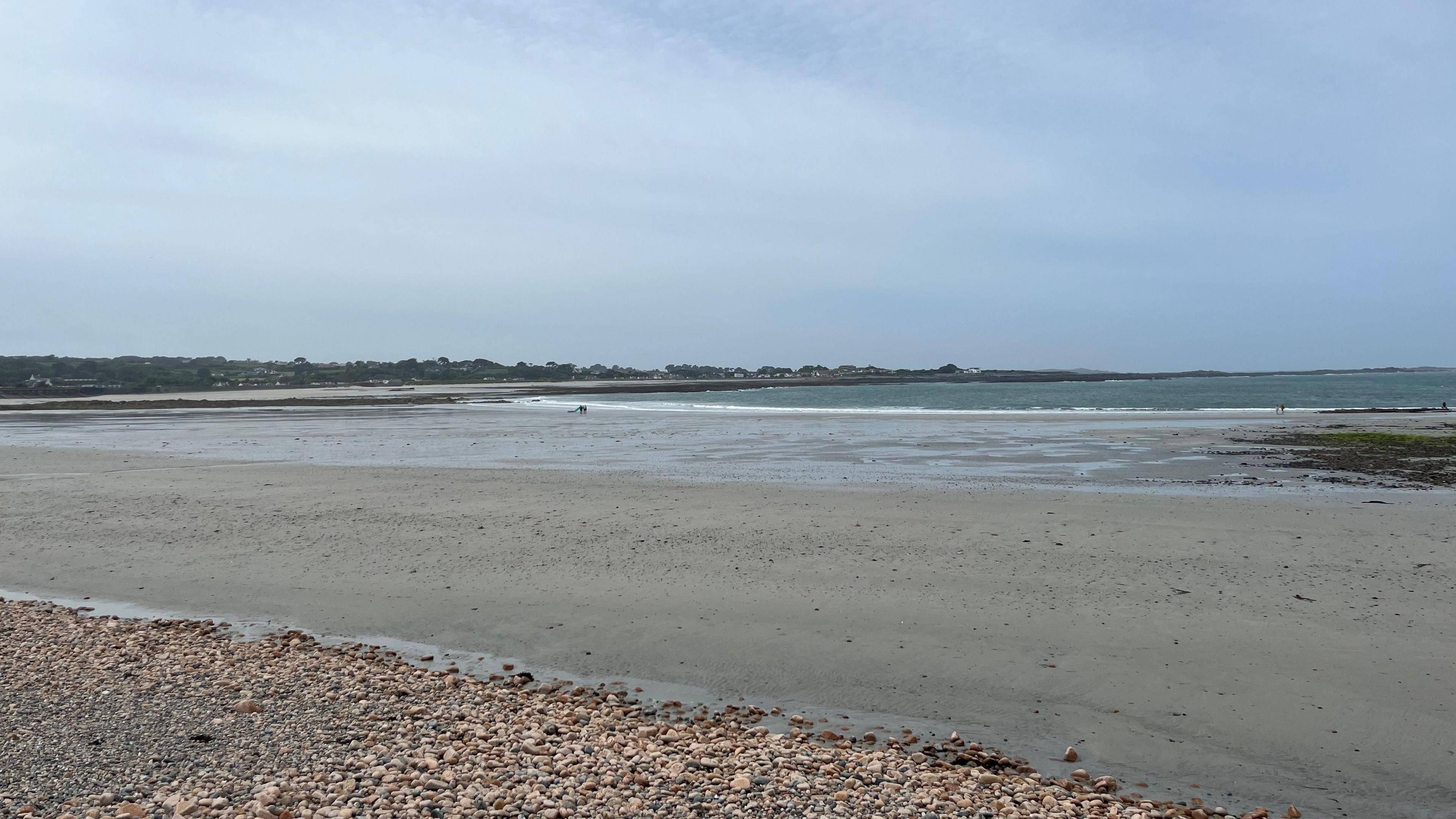 Two people walk on the beach at Vazon Bay on a cloudy day. Part of the sandy beach had water on it from a previous tide.