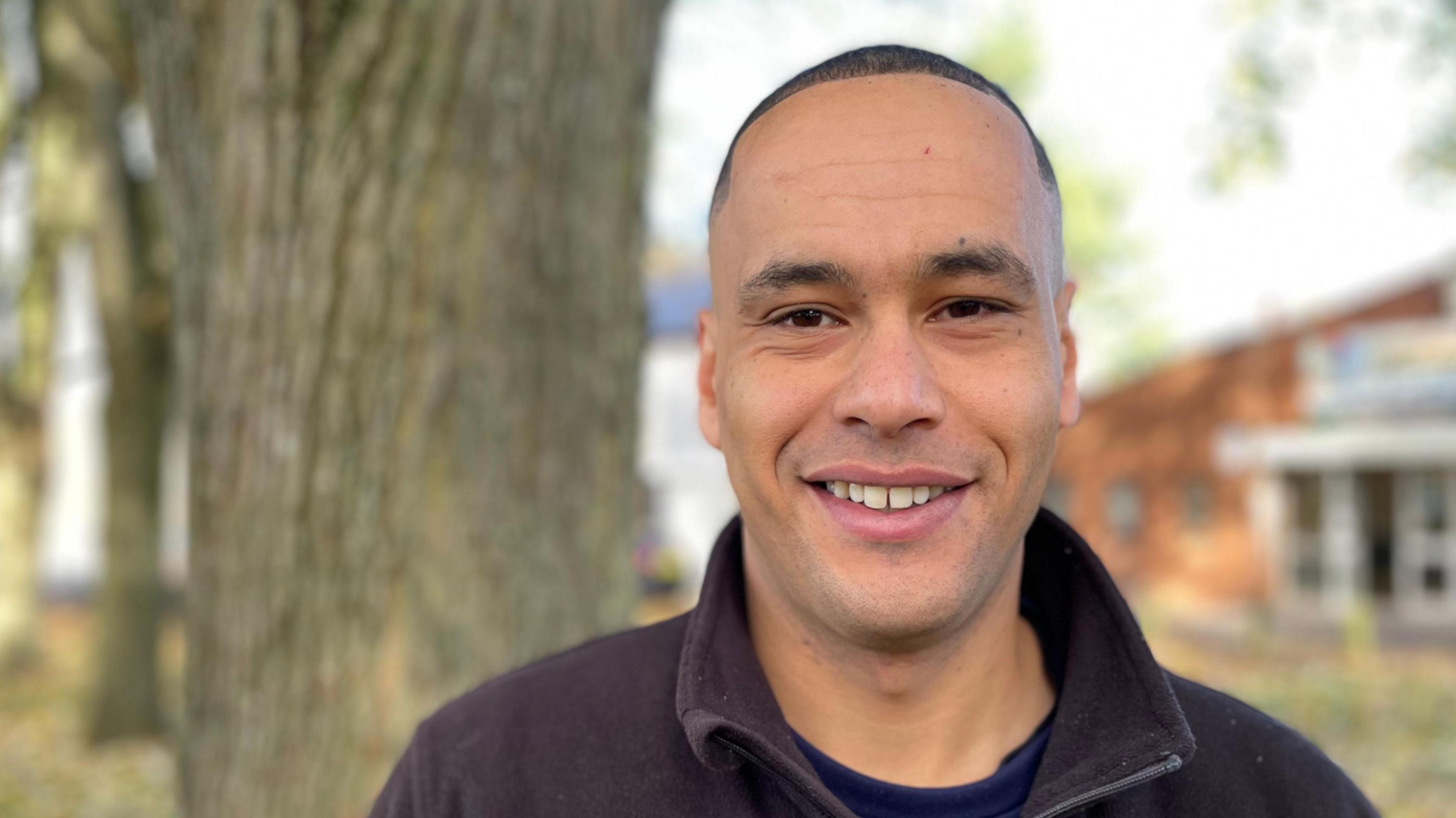 Young man with neatly-cropped hair, smiling, in black top in front of trees.