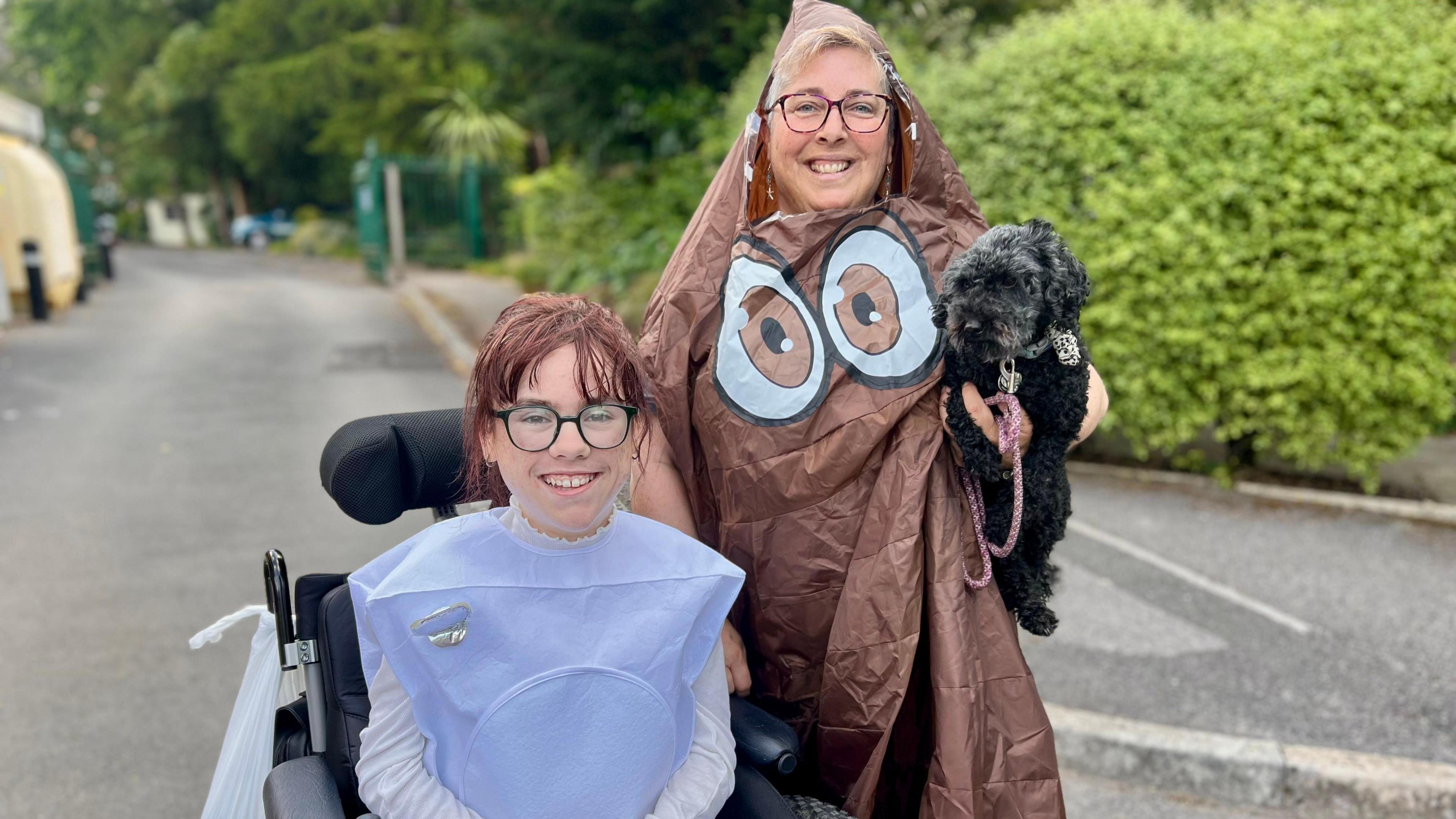 Carmela Chillory-Watson is sittin in a wheelchair dressed as a toilet. She has dark hair which is tied back and is wearing glasses. Her mother, Lucy, is to the left of the image dressed in an emoji poo outfit. She is standing up, holding a black dog under her left arm and is also wearing glasses. The pair are smiling at the camera.