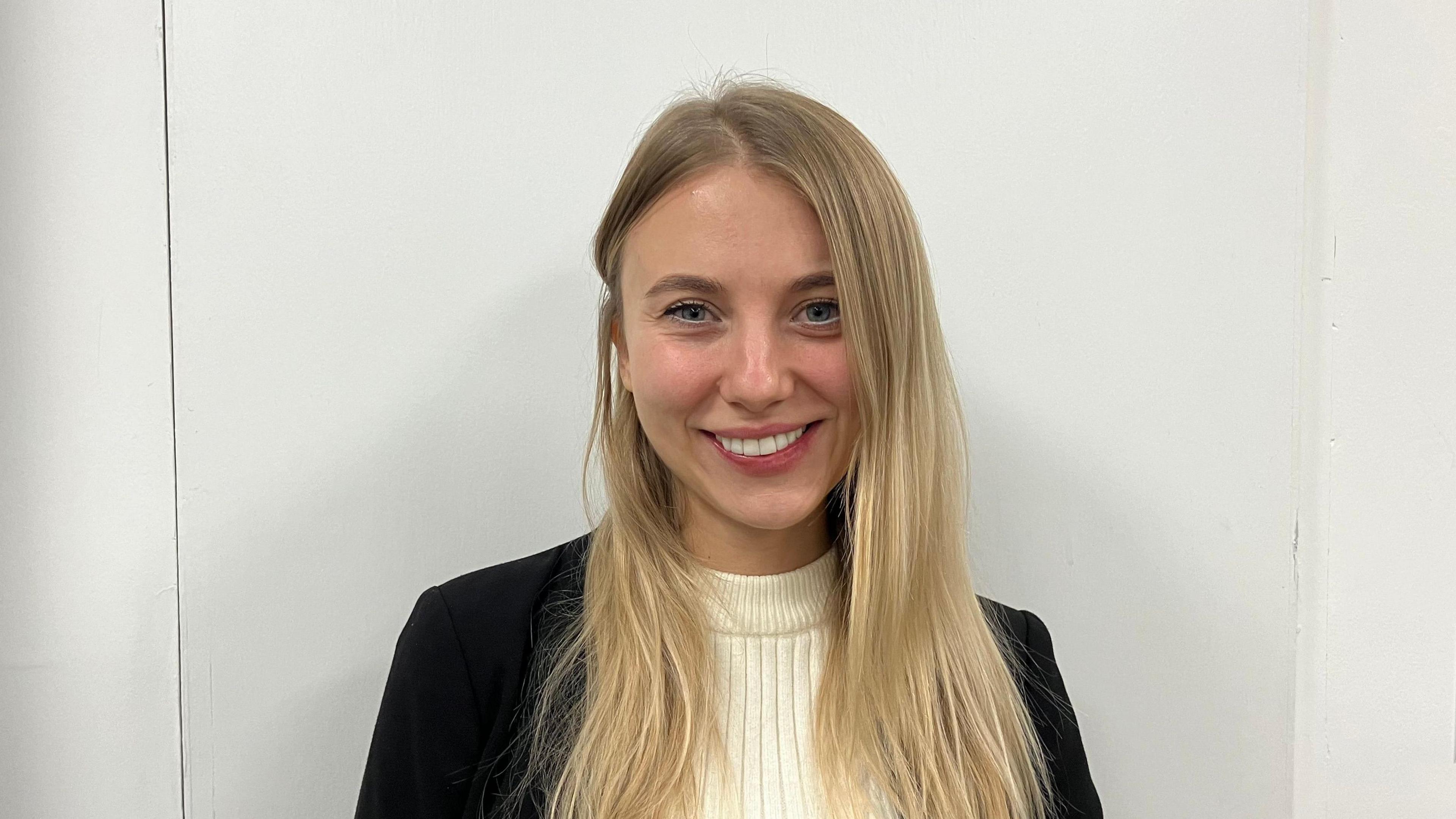 A head and shoulders picture of Anna Sahan standing in front of a white wall. She has long blonde hair and is smiling.