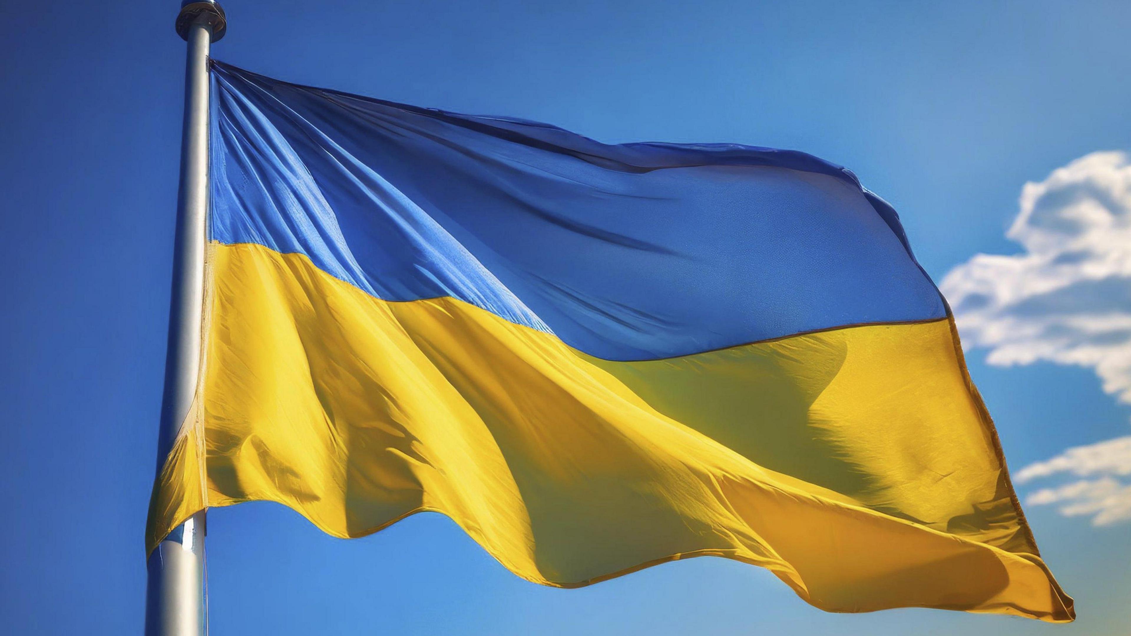 A photo of a Ukrainian flag, with a blue upper half and a yellow bottom half flying on a metal flagpole with a blue sky in the background.