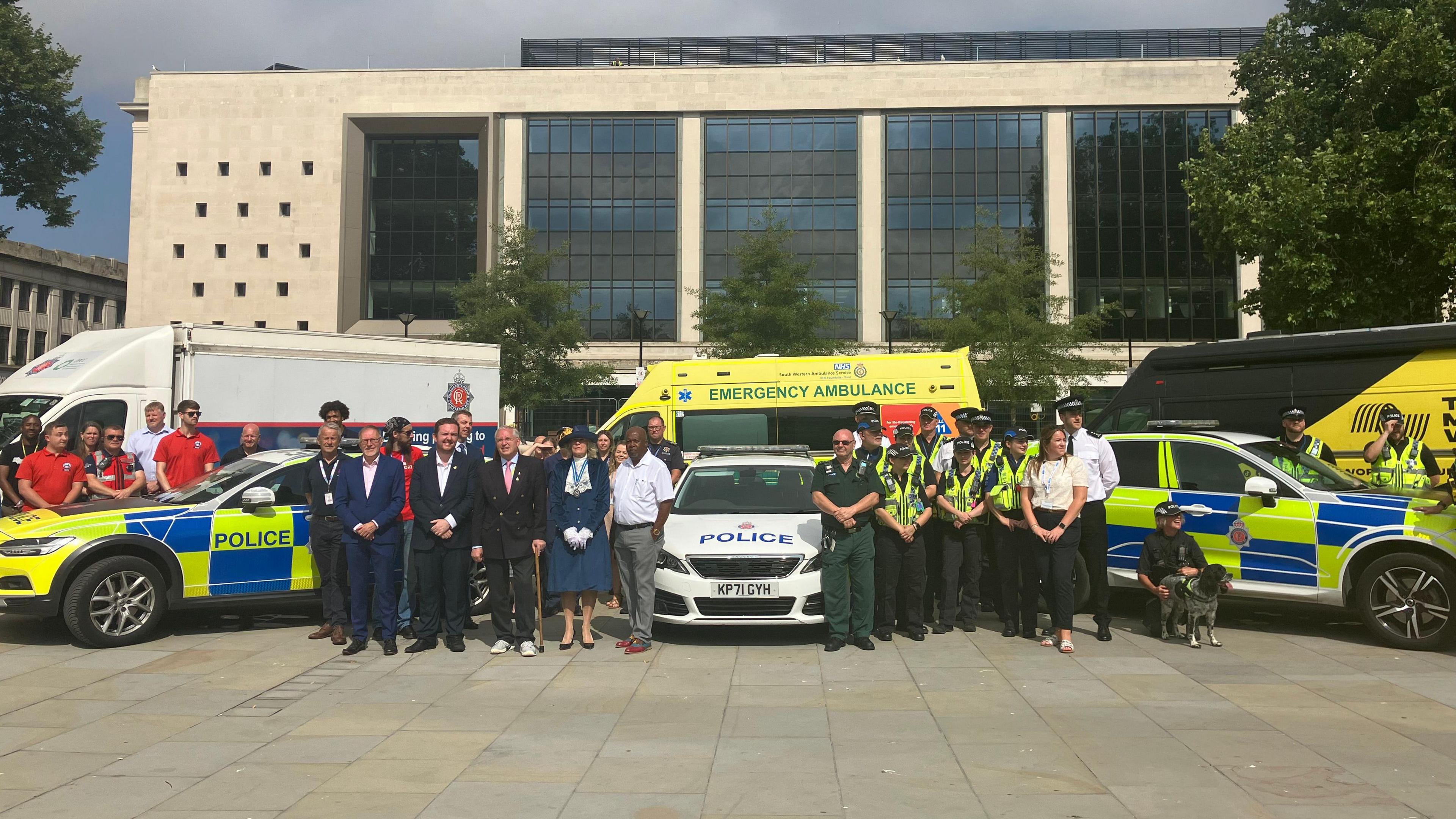 A group of people from various organisations - including police, ambulance, and fire, gather in uniform outside a large, modern building in a city centre square.