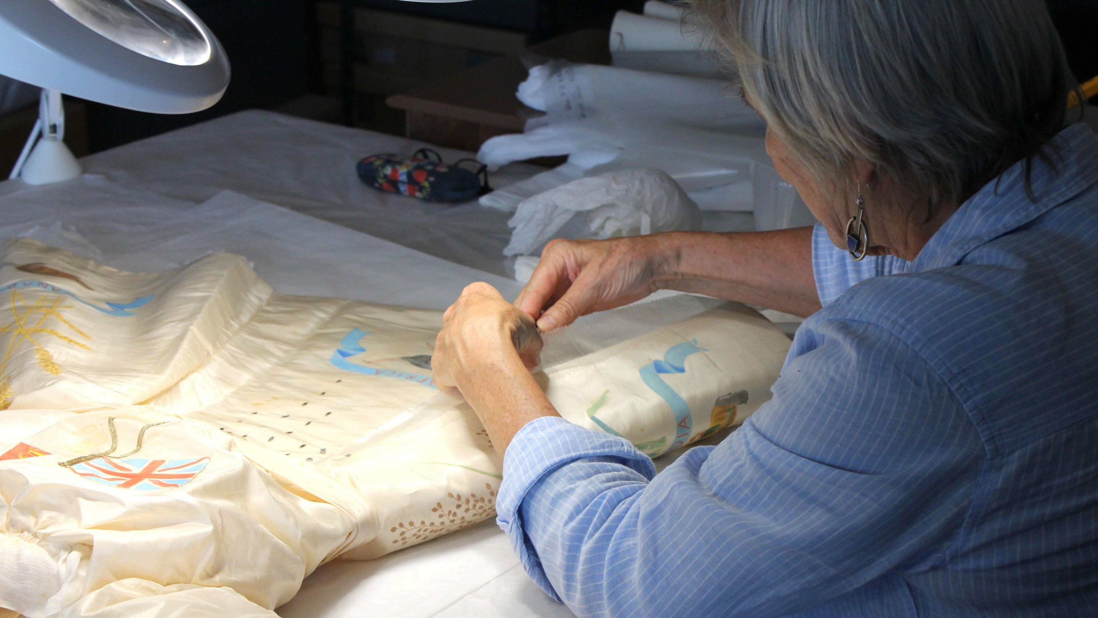 A white silk dress from the 20th Century which is being restored. On the dress are emblems of flags, wheat stems and ribbons. A lady is restoring the dress and she is wearing a blue and white striped shirt with the sleeves rolled up. She has grey hair and her face cannot be seen as she is looking away from the camera and the photograph has been taken over her shoulder.