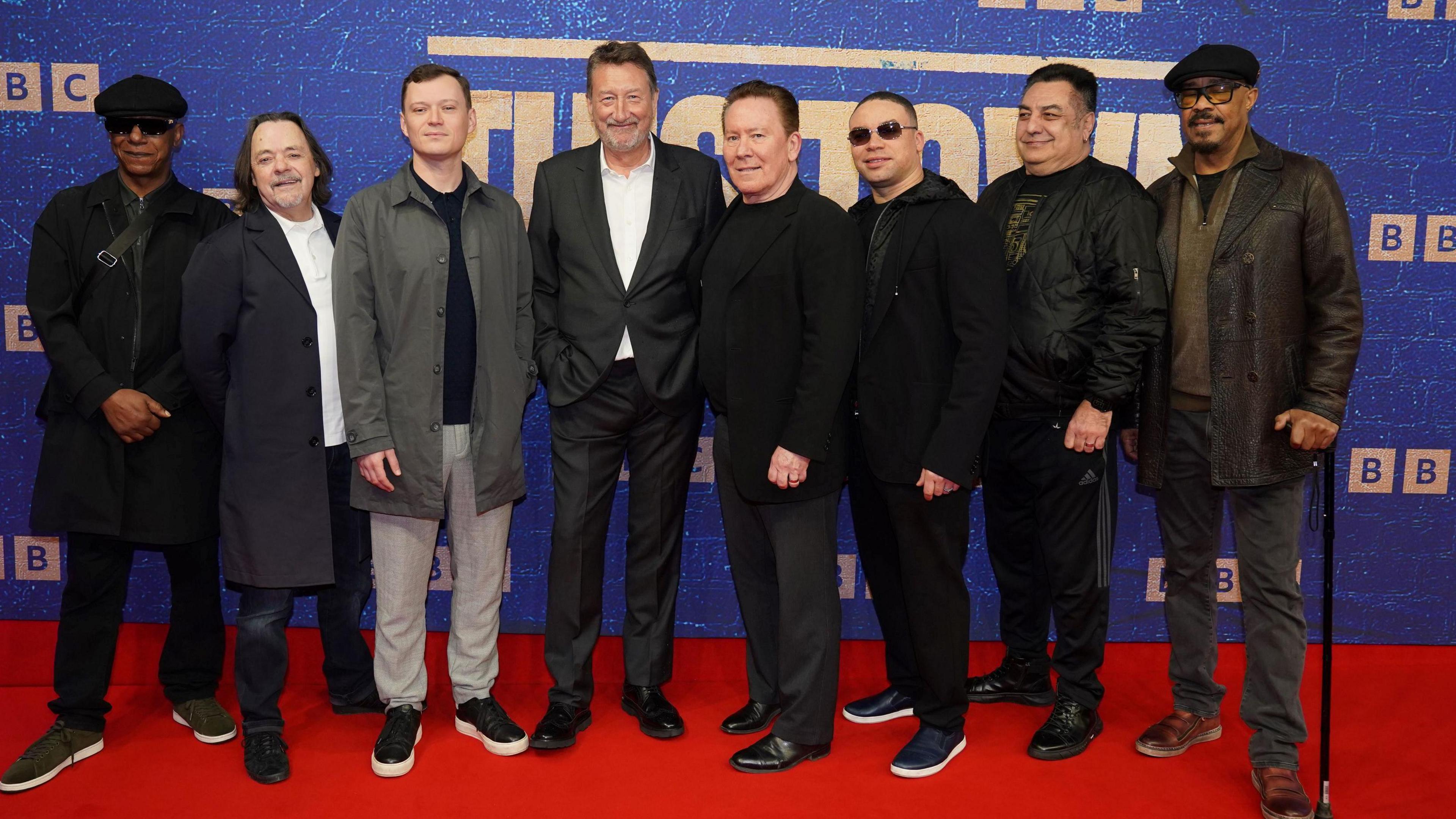 Steven Knight (centre left), with members of UB40 in March 2024. Eight men stand in a line wearing dark clothes. Knight and one UB40 band member have white shirts on.