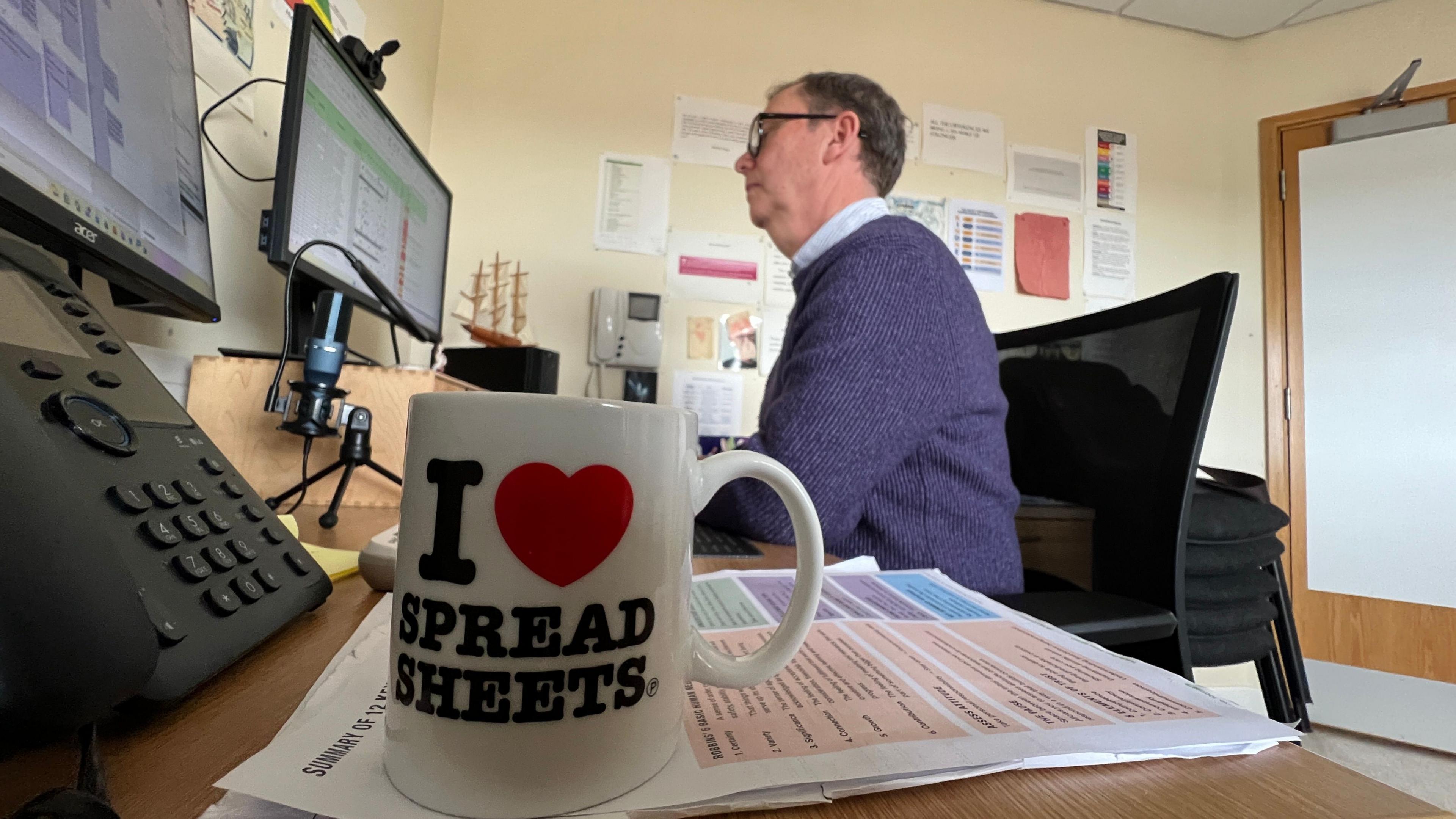 In the foreground is a white mug with the word I love spread sheets on it. The heart is a red heart symbol.  In the background a middle aged man in a blue jumper is at work on a computer.