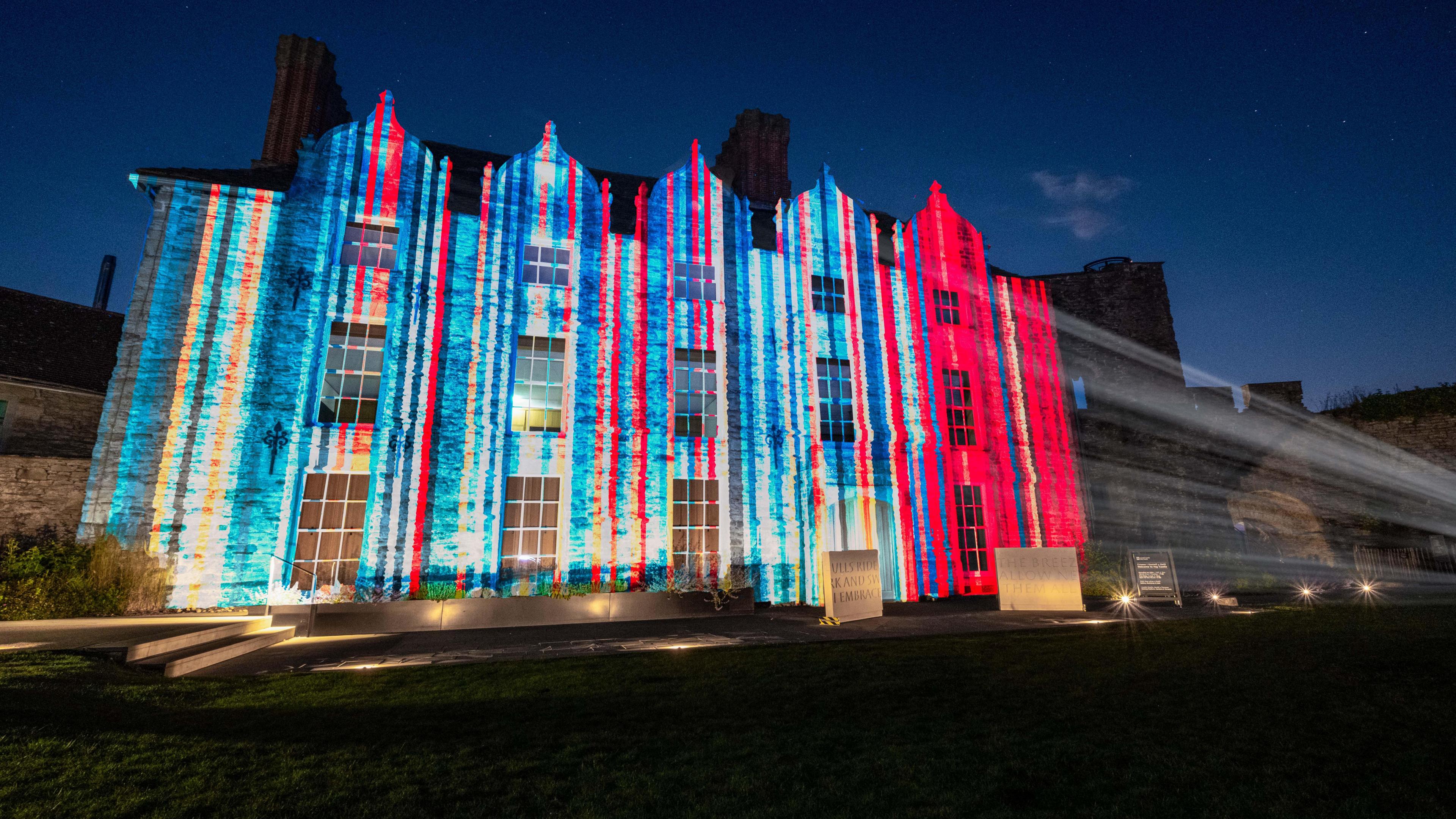 The graphic showing the stripes, starting blue on the left hand side and changing to deep red on the right hand side, being projected onto the stone wall of a castle at night