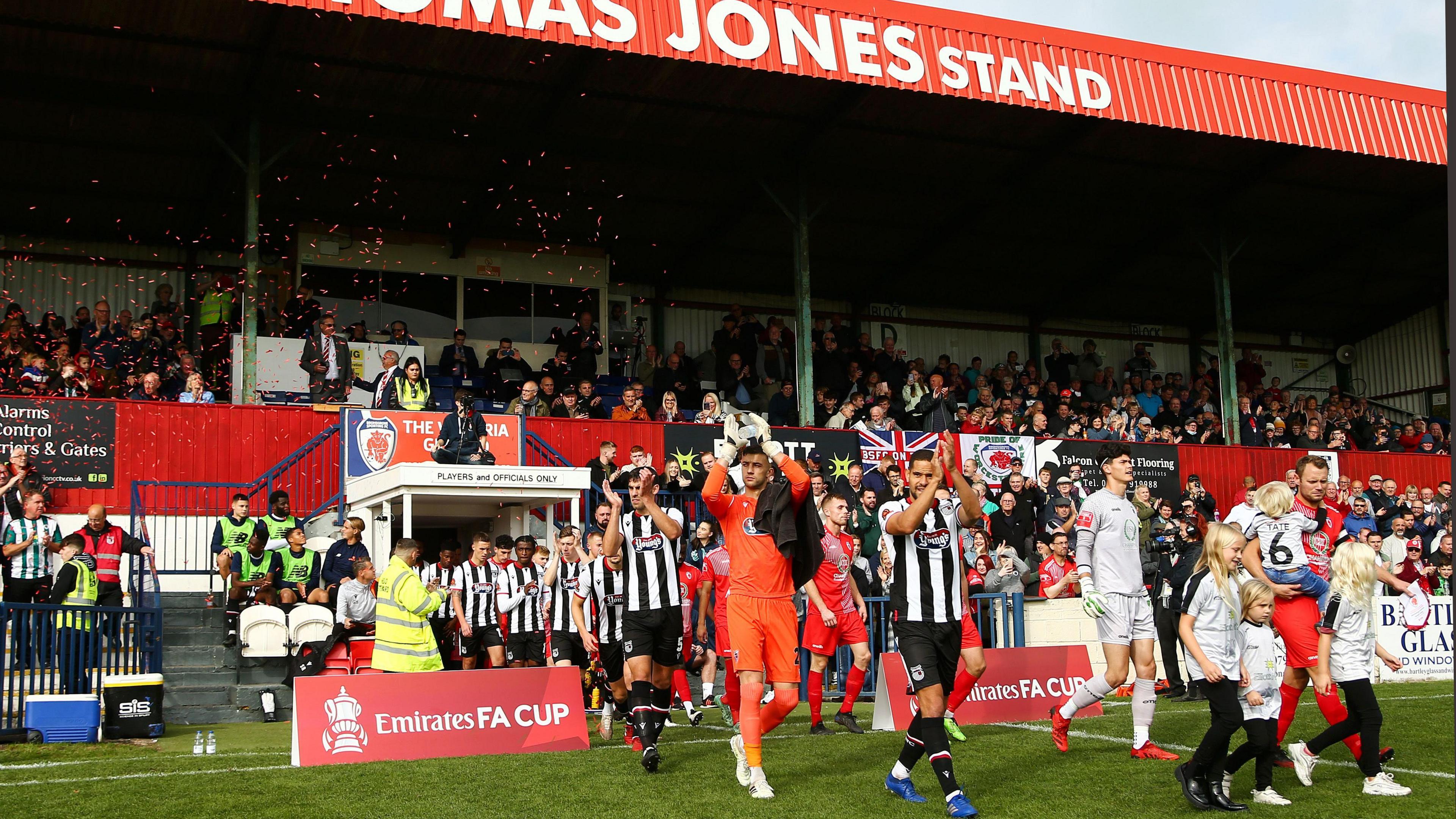 Two football teams, one in black and white stripes and another in all red walk out onto a green pitch from a tunnel underneath a small football that has been painted red and is filled with fans.