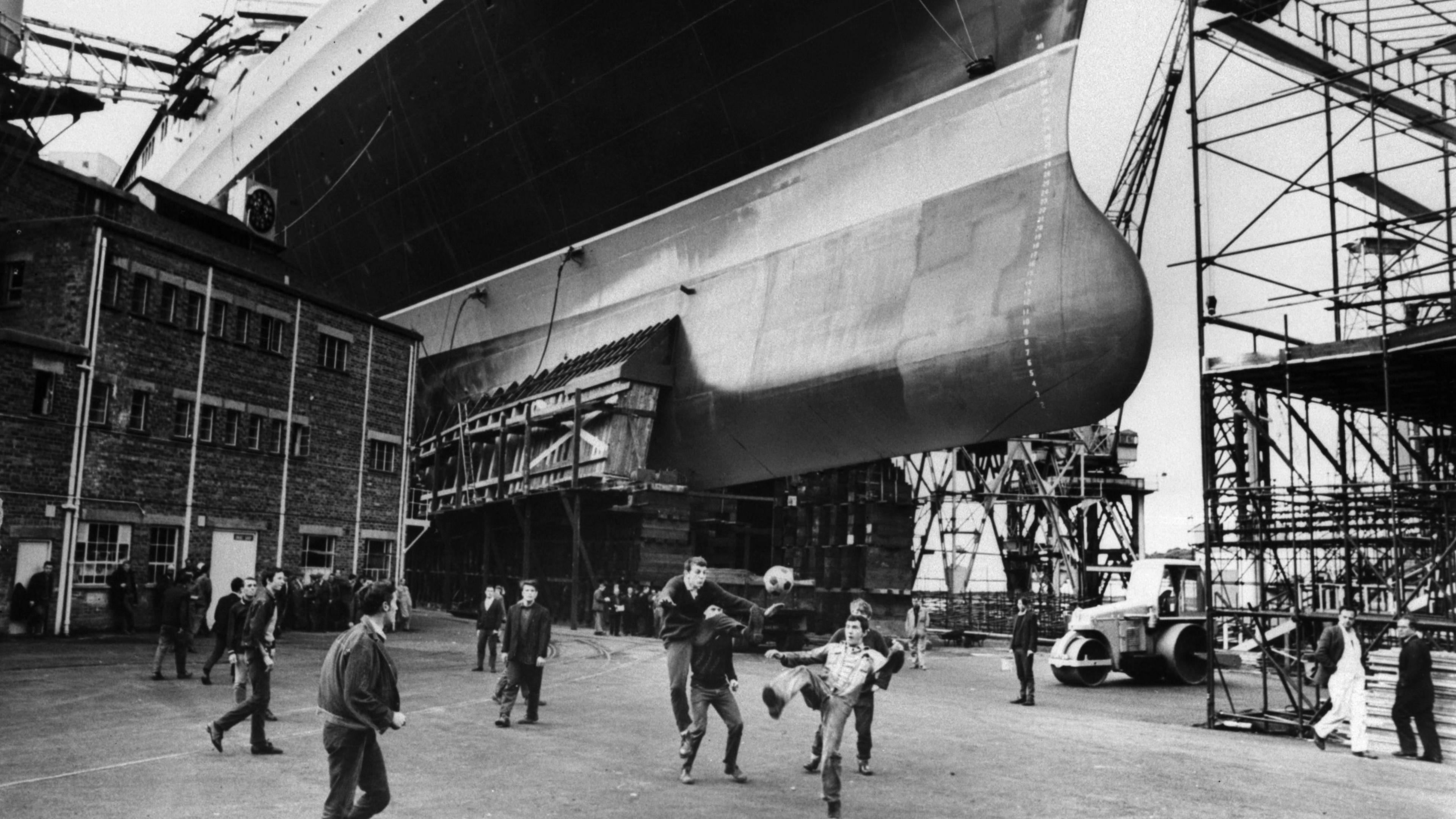 A group of men play football on the dockyard near the hull of a cruise ship being built