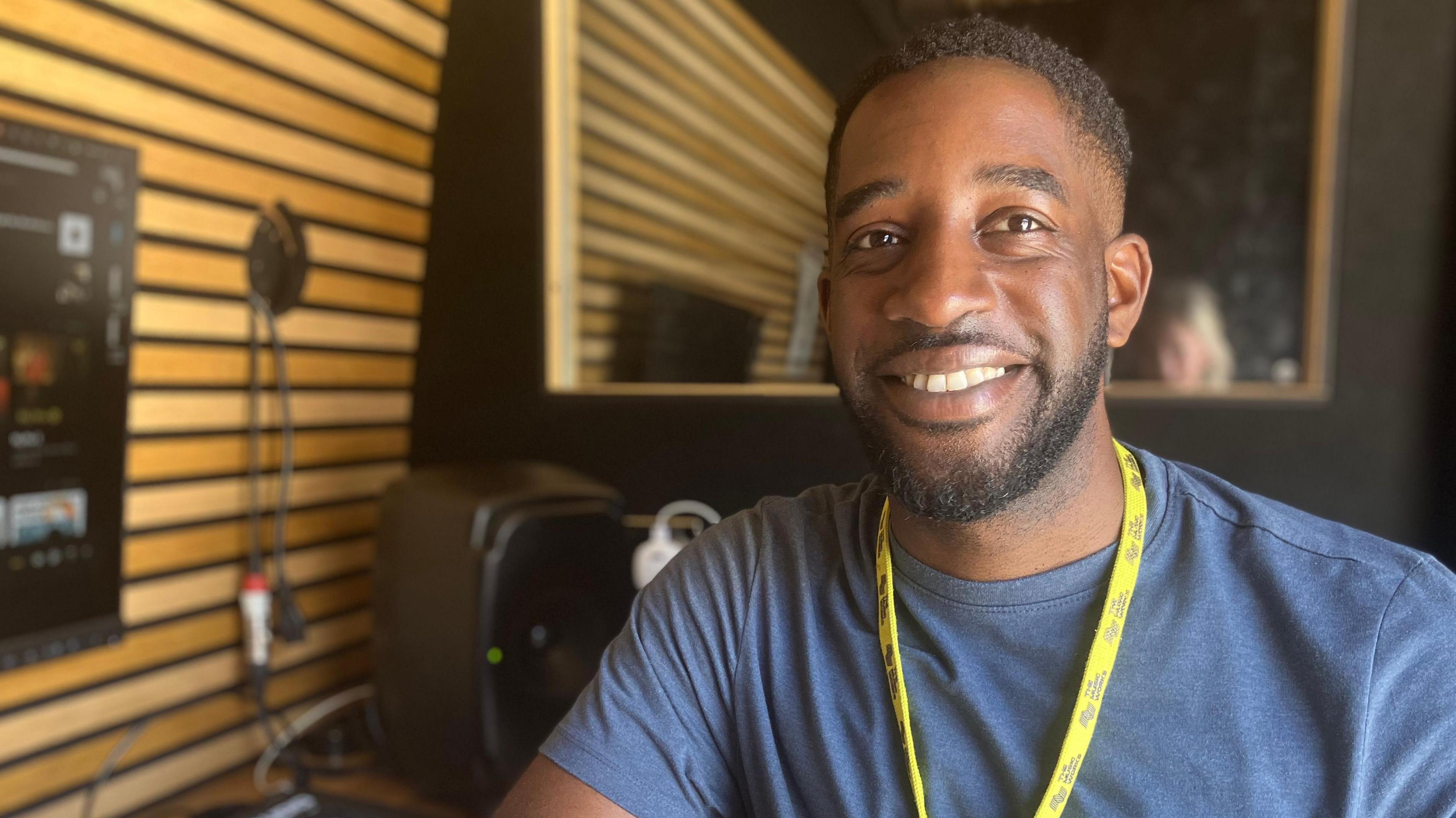 A man smiles as he sits in a wood panelled music studio. He is wearing a blue top and a blue T-shirt. He has short black hair.