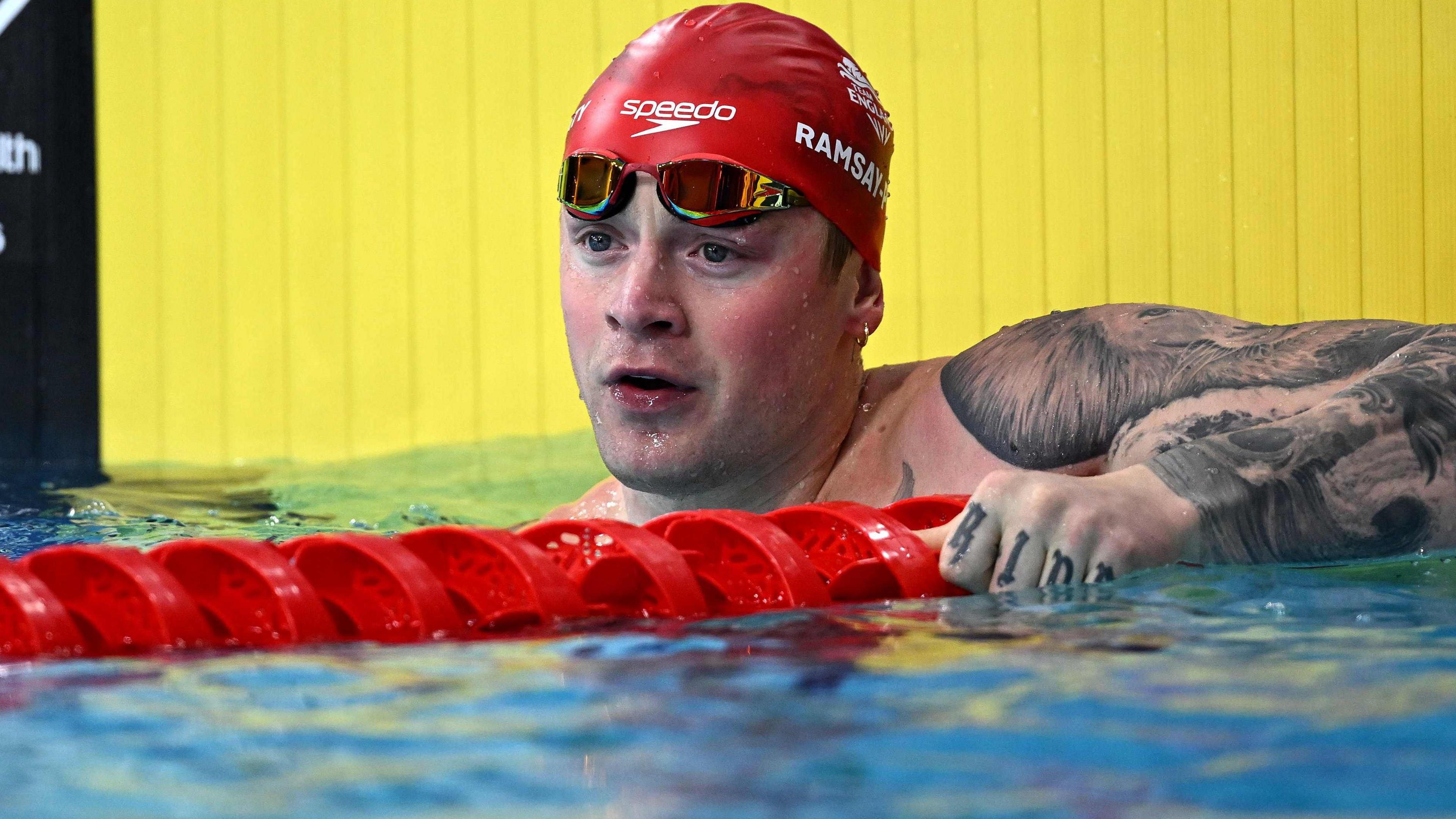 Adam Ramsay-Peaty, wearing a red Speedo swimming cap and goggles, holds the red lane separator in the pool after the 50m breaststroke heats at the 2026 Commonwealth Games