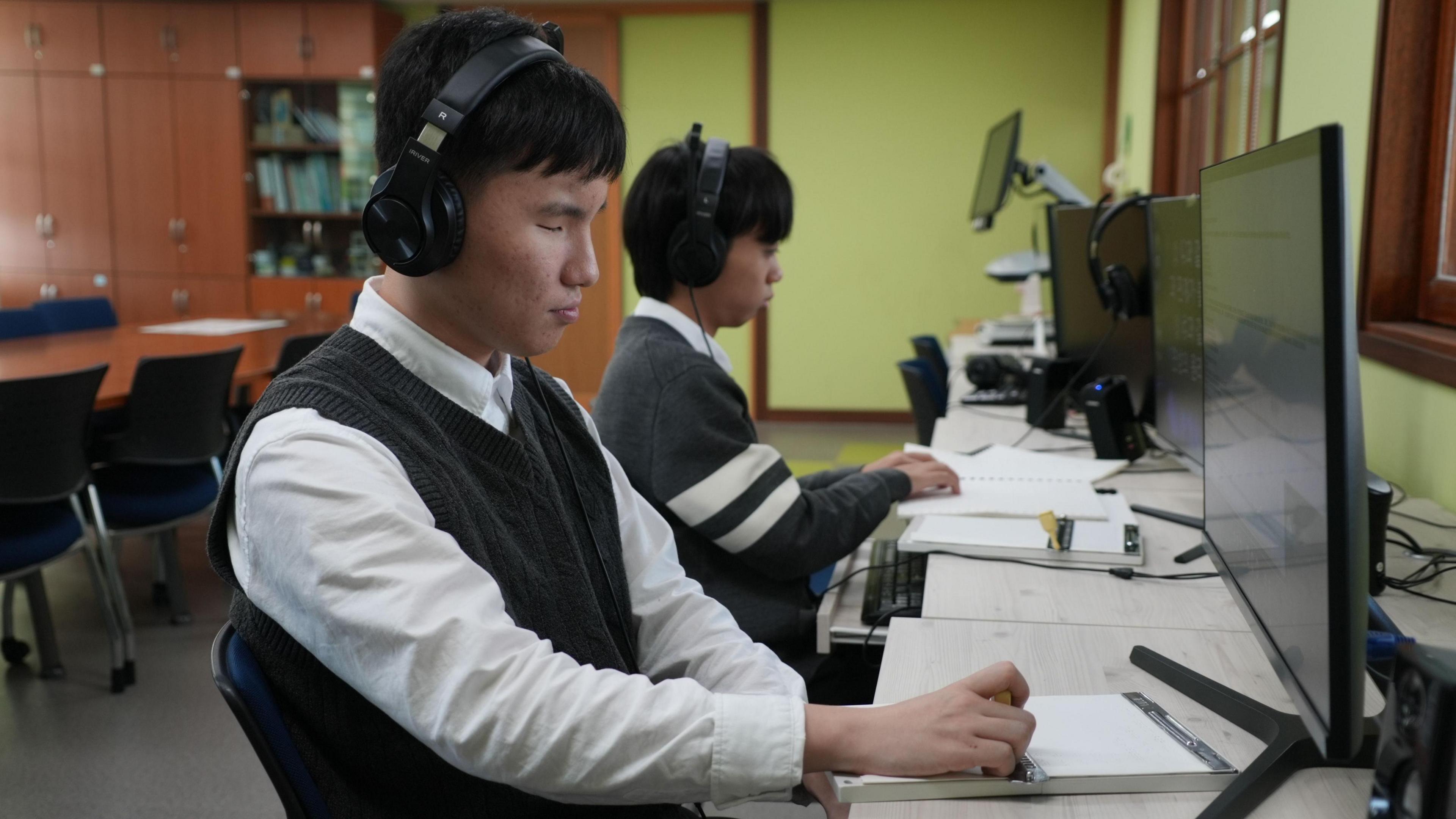 Students wearing over-ear headphone sit at desks in front of computers screens and use screen readers and braille