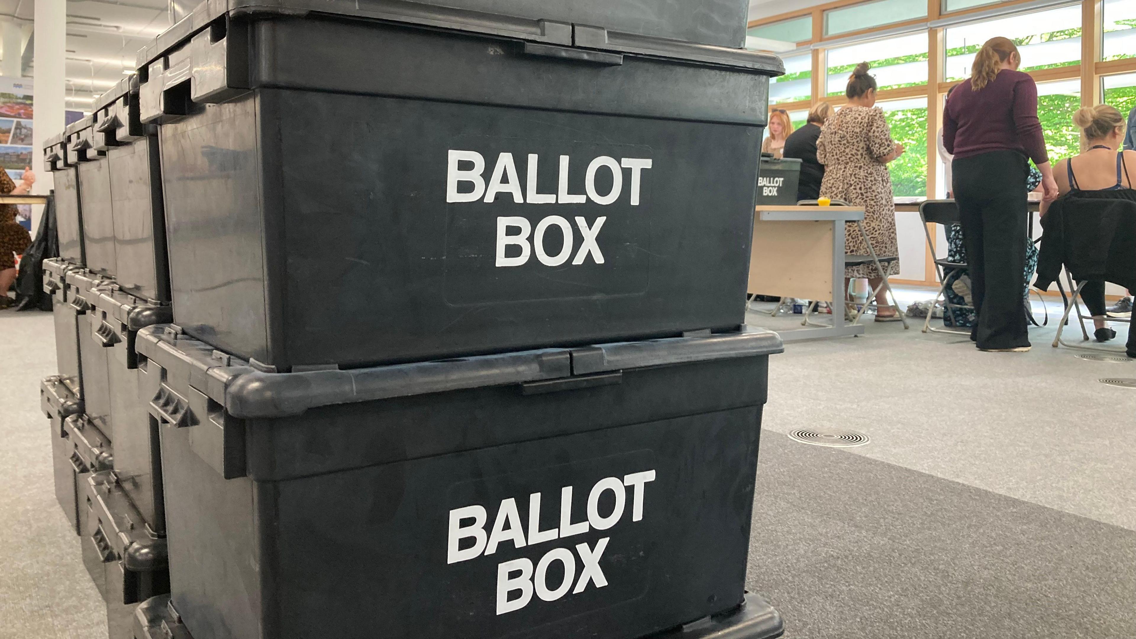 There are a pile of black ballot boxes stacked inside a counting centre. A number of people can be seen in the background sitting and standing at desks.