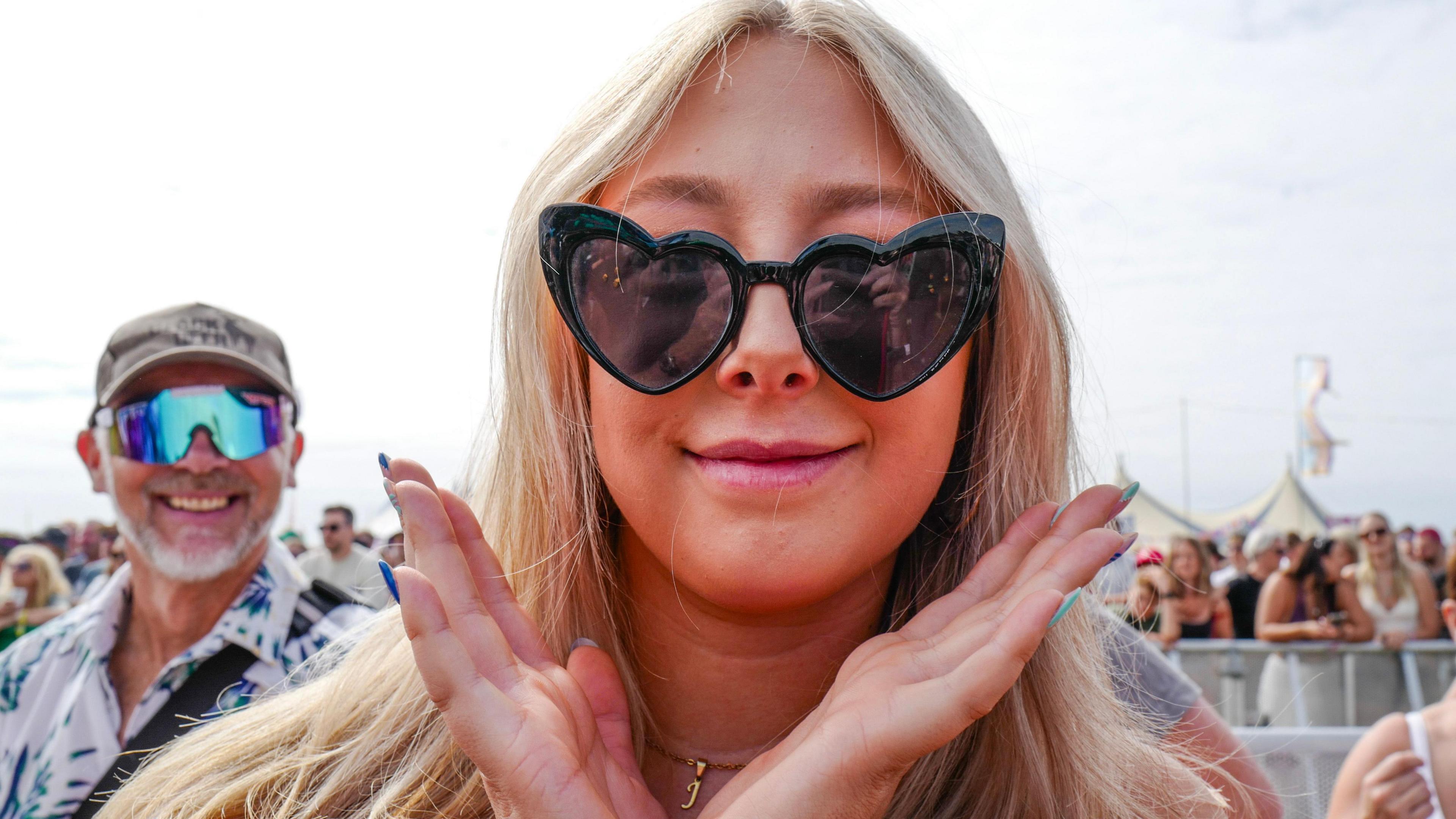 A woman with long blonde hair and black heart-shaped sunglasses holds both hands up near her face. In the background, there is a domed tent and other festivalgoers.