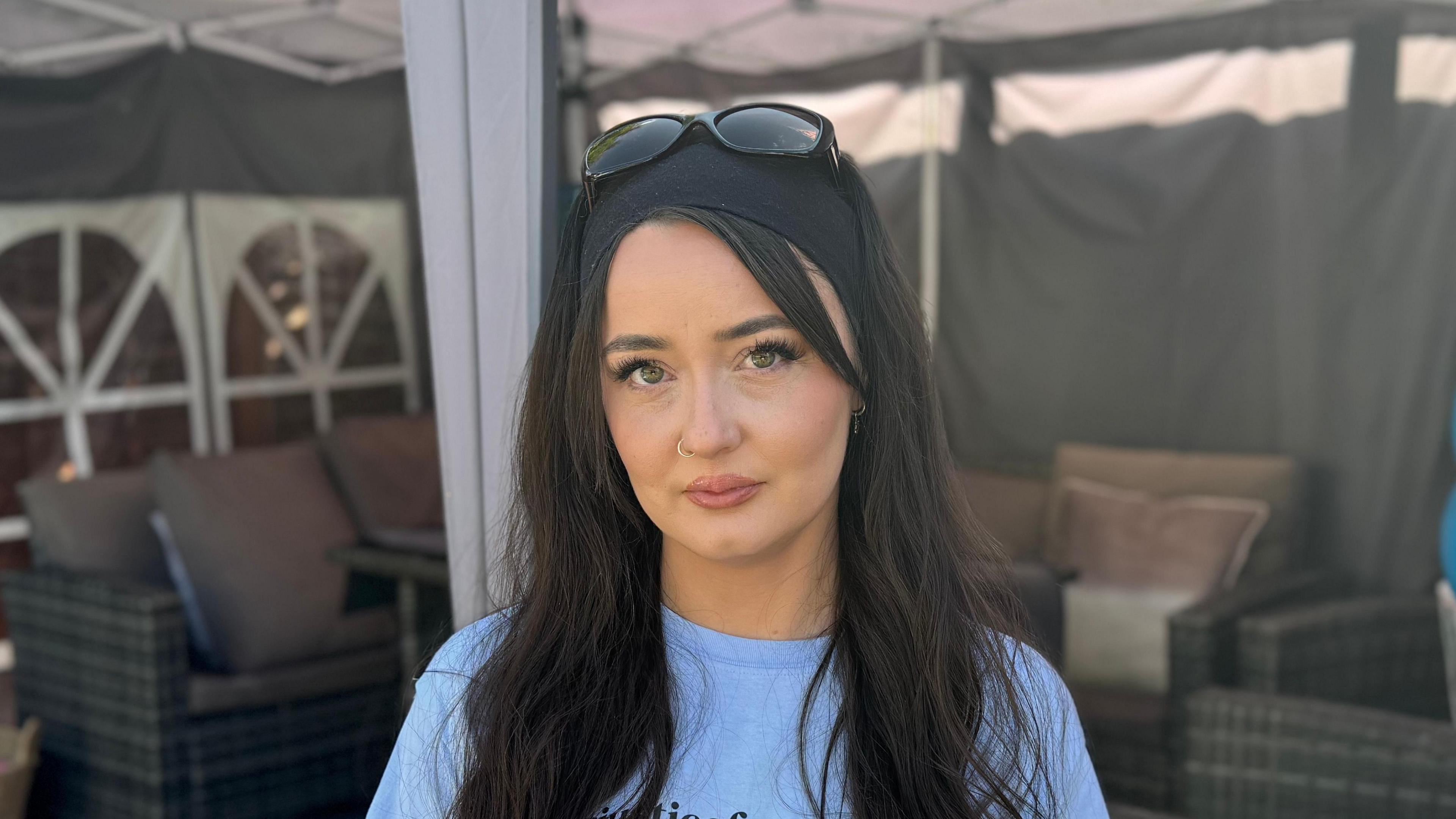 A head and shoulders image of Charleigh Mitchell. She has long black hair and is looking into the camera. She is standing in a garden and wearing a light blue T-shirt. 