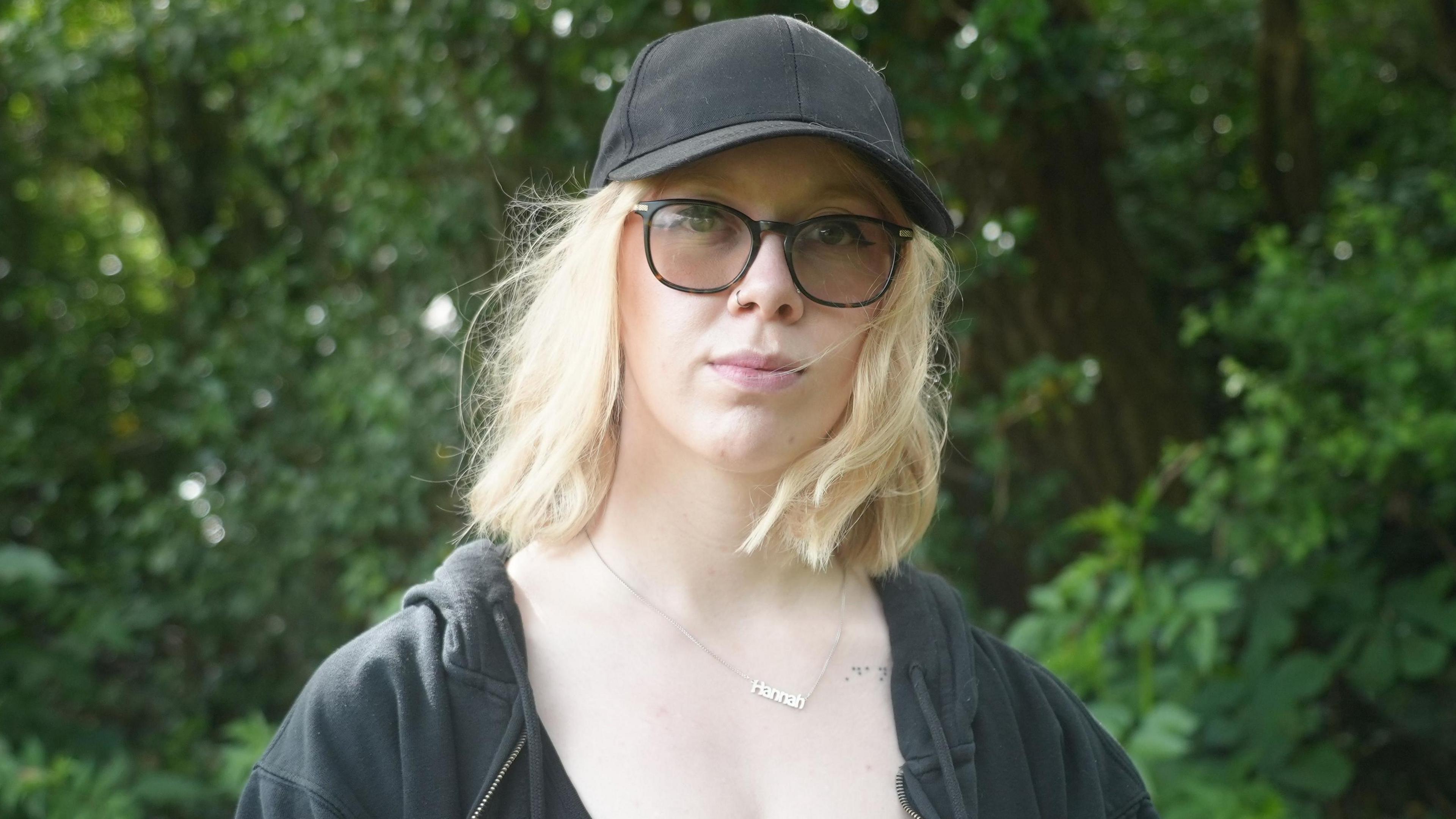 A head and shoulders shot of Hannah Burgess looking at the camera in front of green trees in the background. Shge is wearing a black cap and glasses.