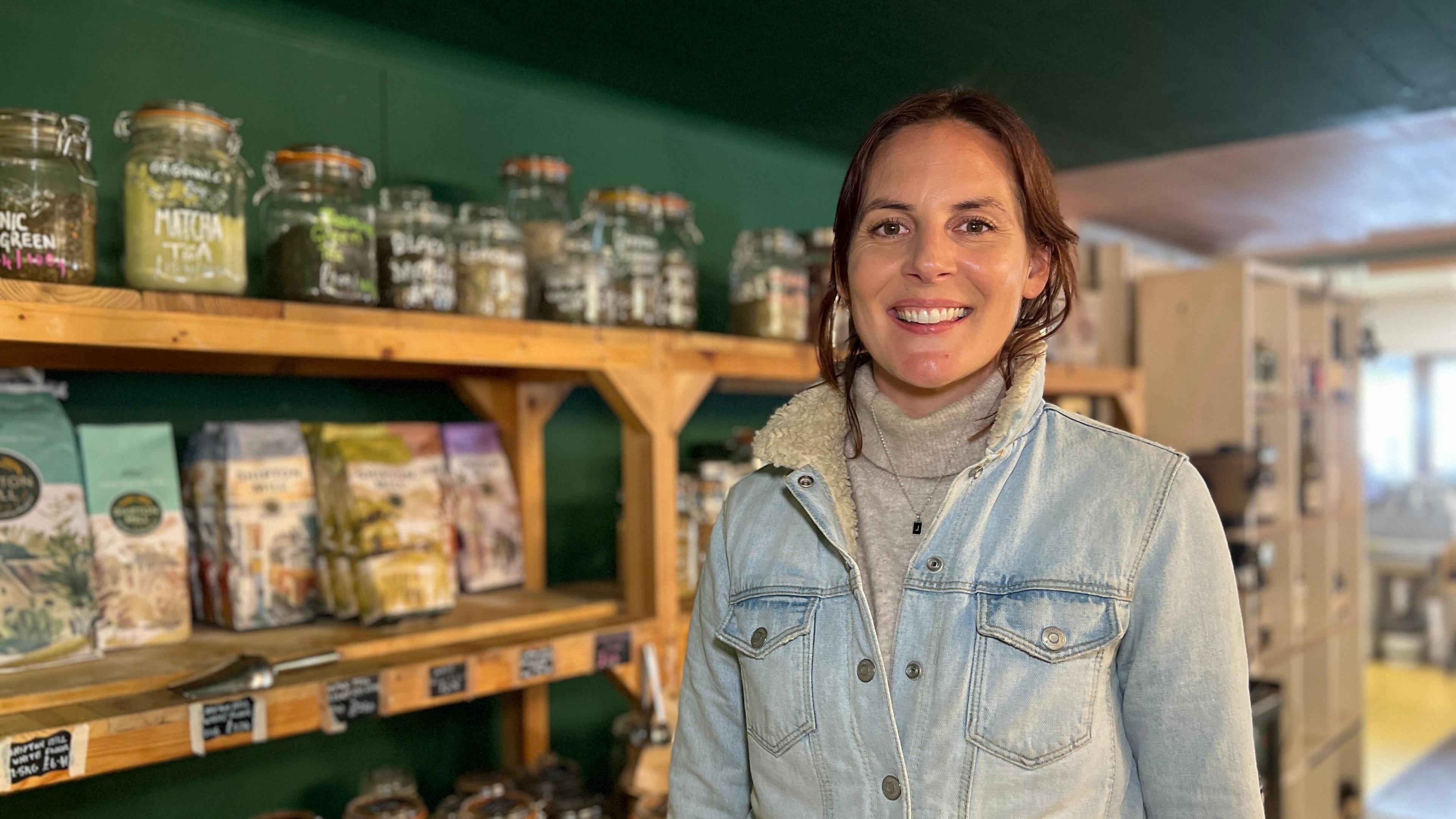 Jess stands in front of shelves filled with various jars and packages in a store. Shel is wearing a light denim jacket over a turtleneck sweater. The shelves behind them contain glass jars labeled with different types of tea and other items, as well as some packaged goods. The background shows more shelving units and the interior of the store.