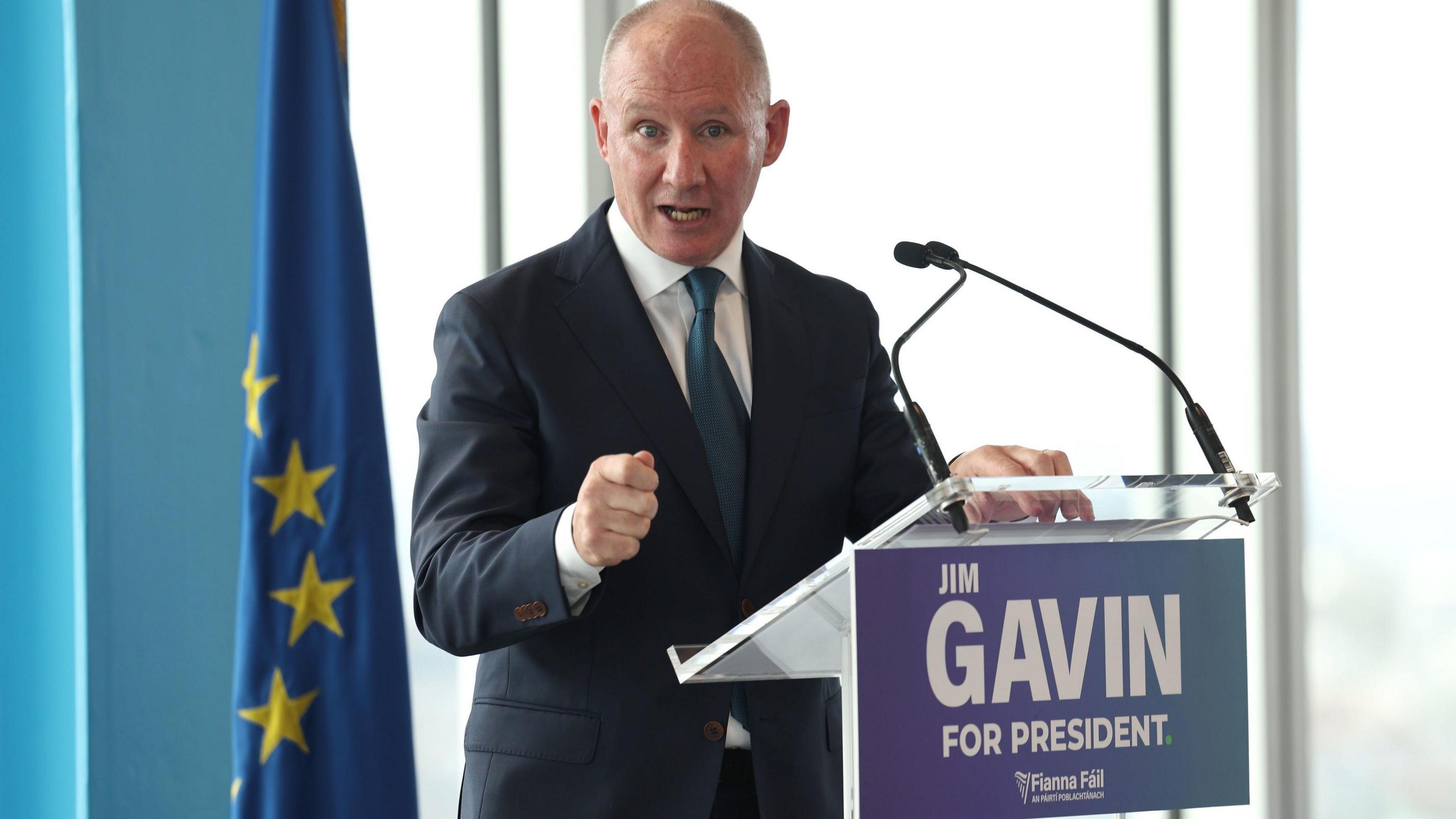 Jim Gavin has receding hair. He is wearing a dark suit and a green tie and pointing towards the camera.
