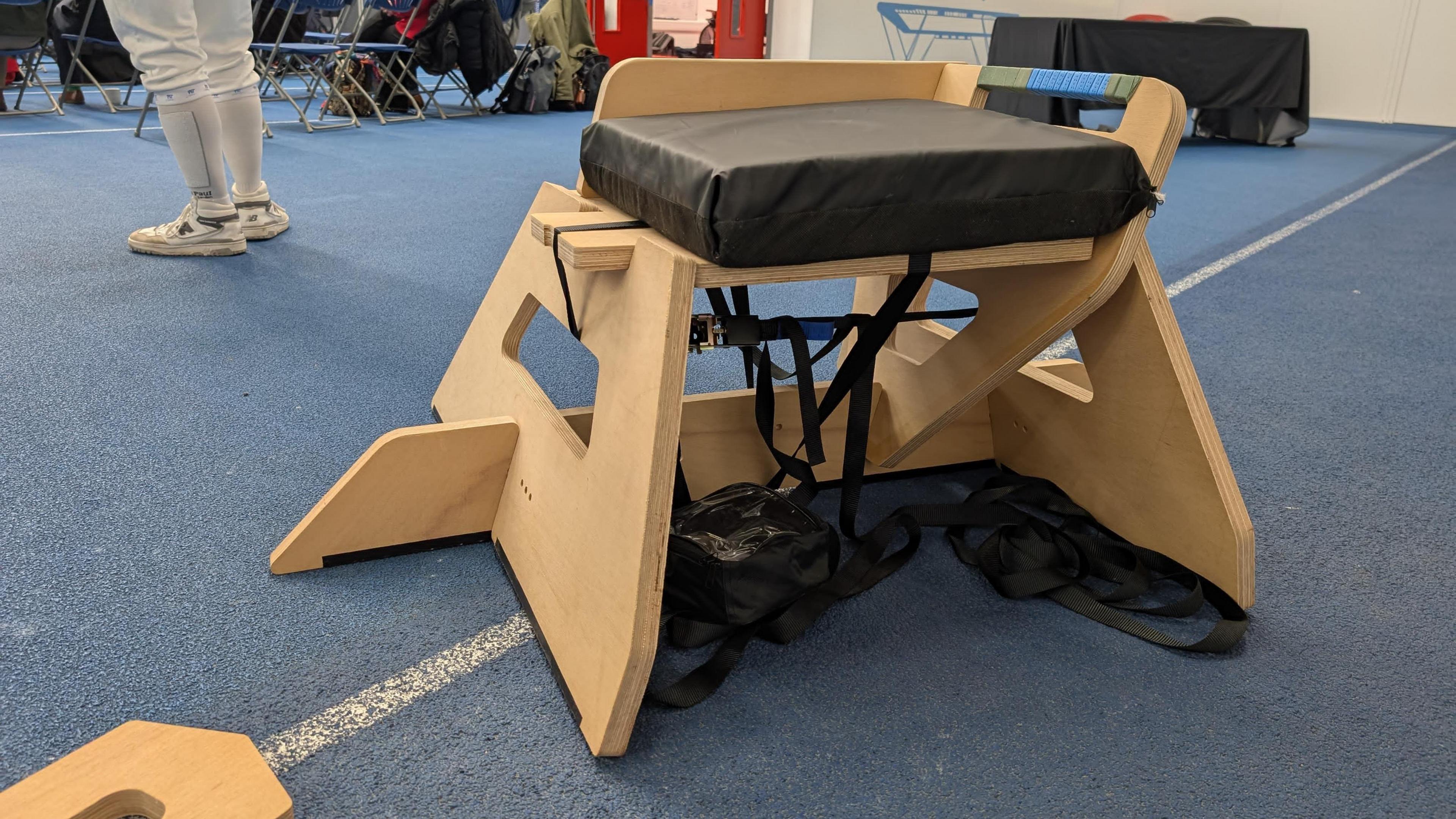 A frame made of plywood with a black cushion on top is pictured on the floor of a sports centre.