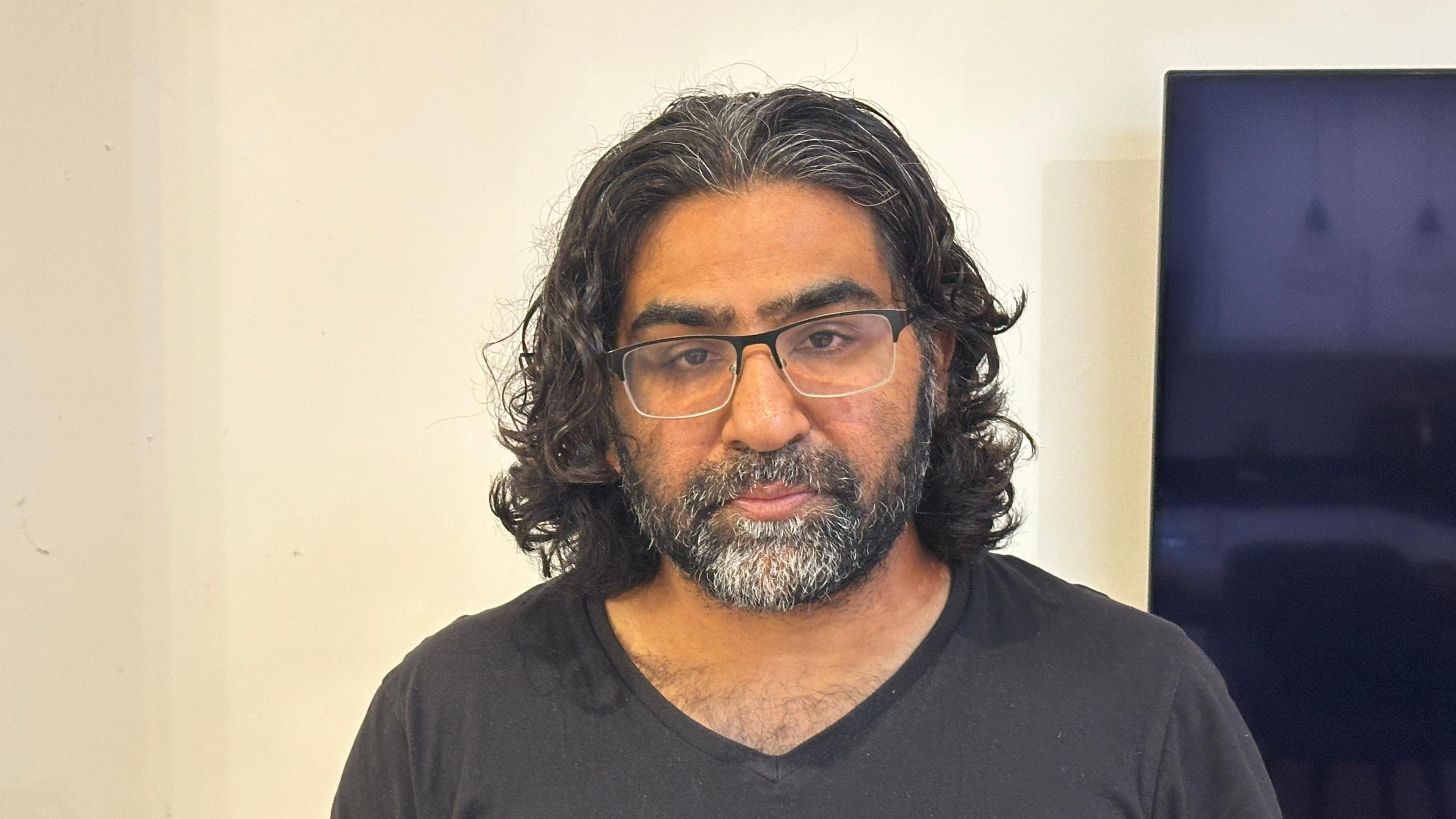 Mohammad Aslam looks to the camera. He has shoulder length dark wavy hair and black rimmed glasses. He has a faded black V-neck t-shirt on and is stood in front of a cream coloured wall with a television to the right hand side of the image. 