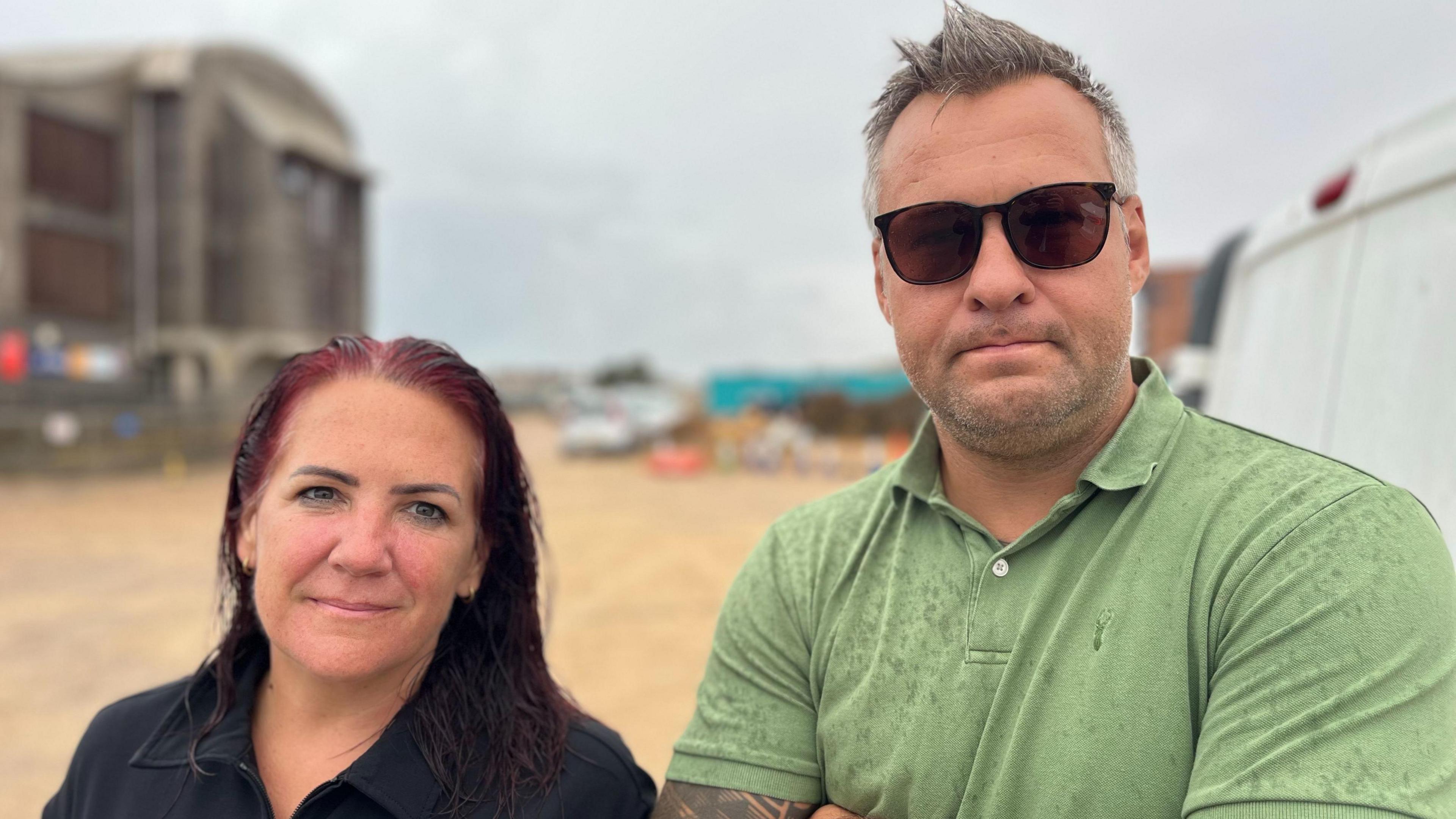 Liz Donnelly and Lee Smith stand on a beach near Shoreham seafront. Donnelly is wearing a dark top, while Smith is wearing sunglasses and a green polo shirt. Buildings and beach structures can be seen in the background.