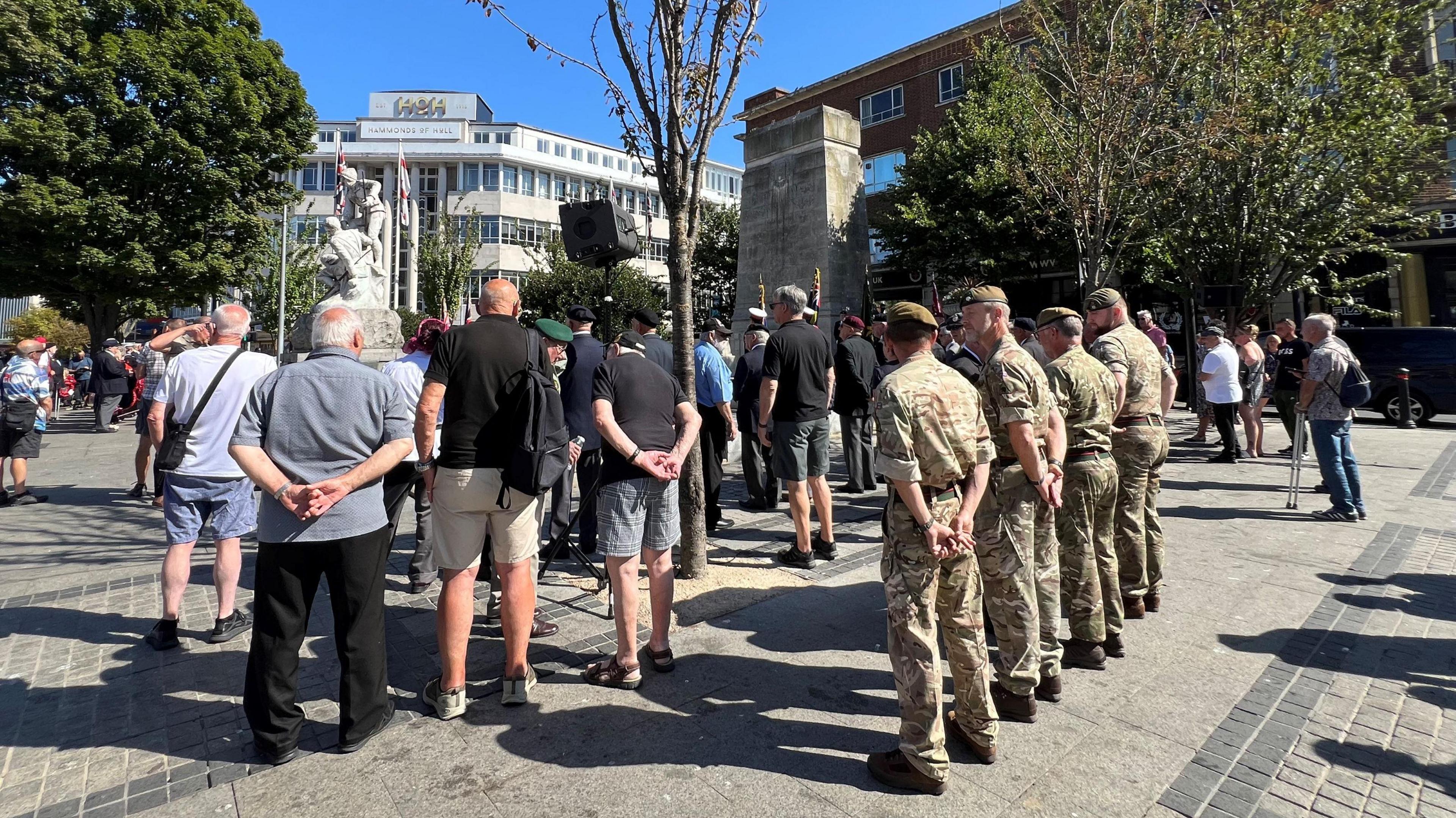 A group of people facing away from the camera and towards a ceremony. Four men at the back of the group are wearing camouflage uniform. A white building is in the distance. The sky is blue.