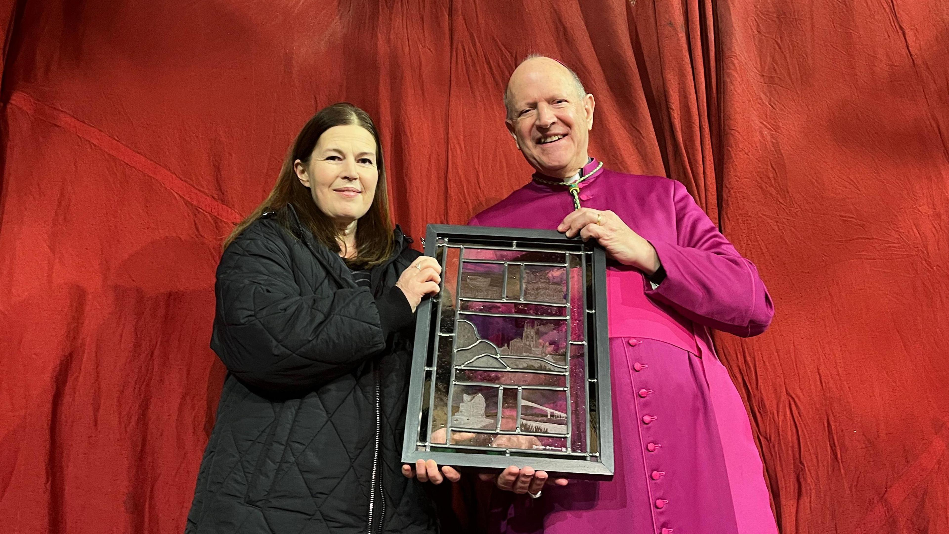 Danielle Hopkinson - a woman wearing a black jacket - presenting the Right Reverend Martin Seeley - a man wearing pink regalia - with a stained glass artwork. They are both standing in front of a red material backdrop 