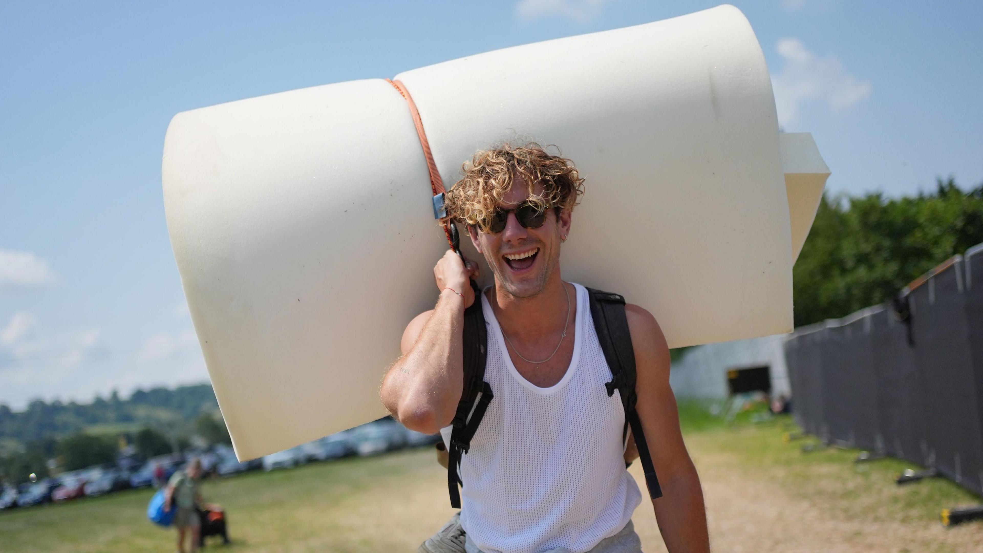 Man in a white vest top and sunglasses carrying a mattress on his back at Glastonbury