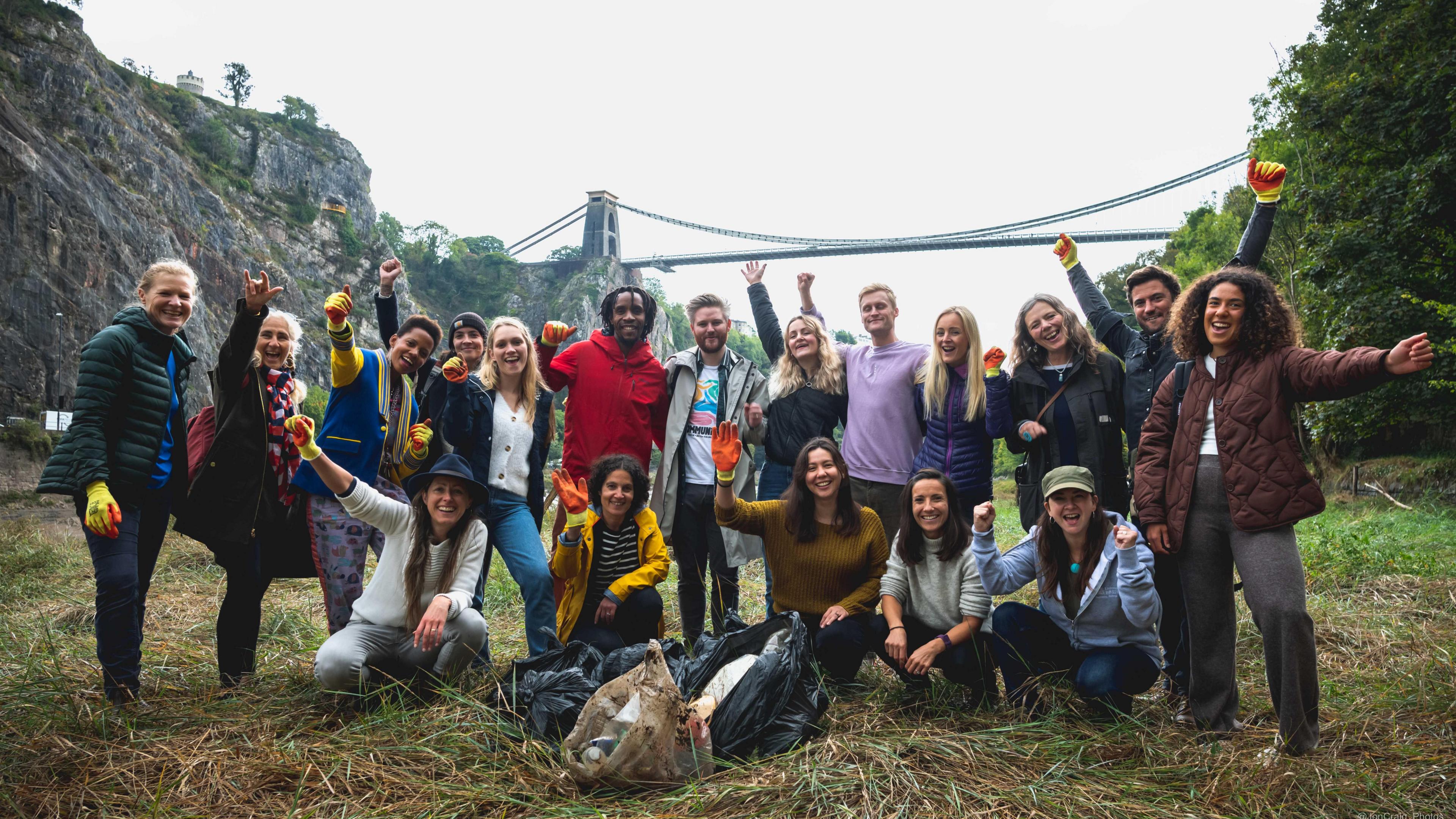 A group of 18 litter pickers - of different ages and genders - stand joyfully with their arms in the air, with the Clifton Suspension Bridge in the background. They are wearing gloves and there are black bin bags on the floor, filled with what appears to be rubbish.