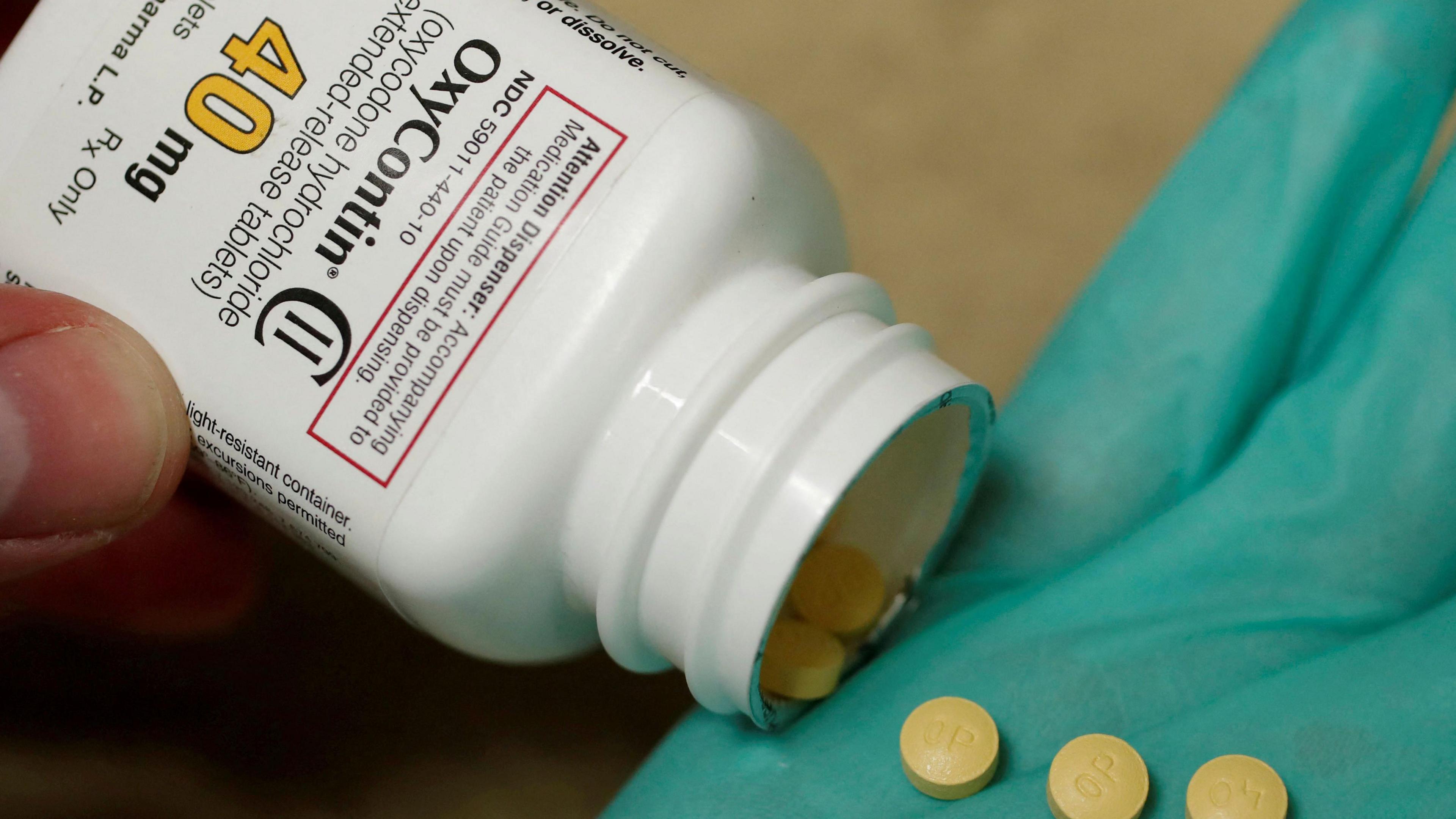 A hand holds a white bottle reading "OxyContin", pouring out small round pills.