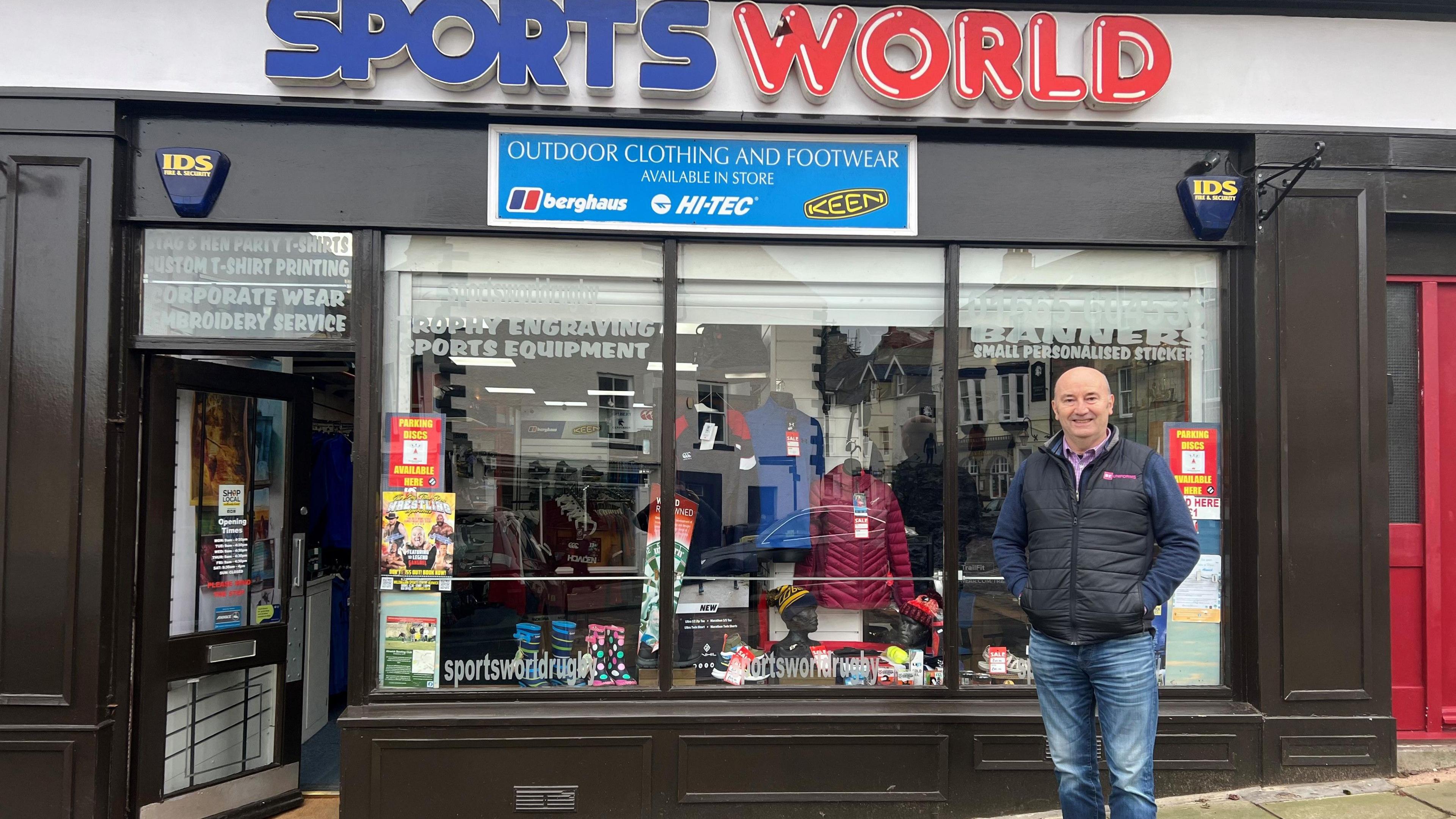 Andrew Hodgson is standing outside his business Sports World. He is a 60-year old-man, bald, wearing a blue top under a black gilet and has blue jeans. Outdoor clothing can be seen in the shop window below the shop logo which consists of the word Sports in blue letters and World in red with white shading.