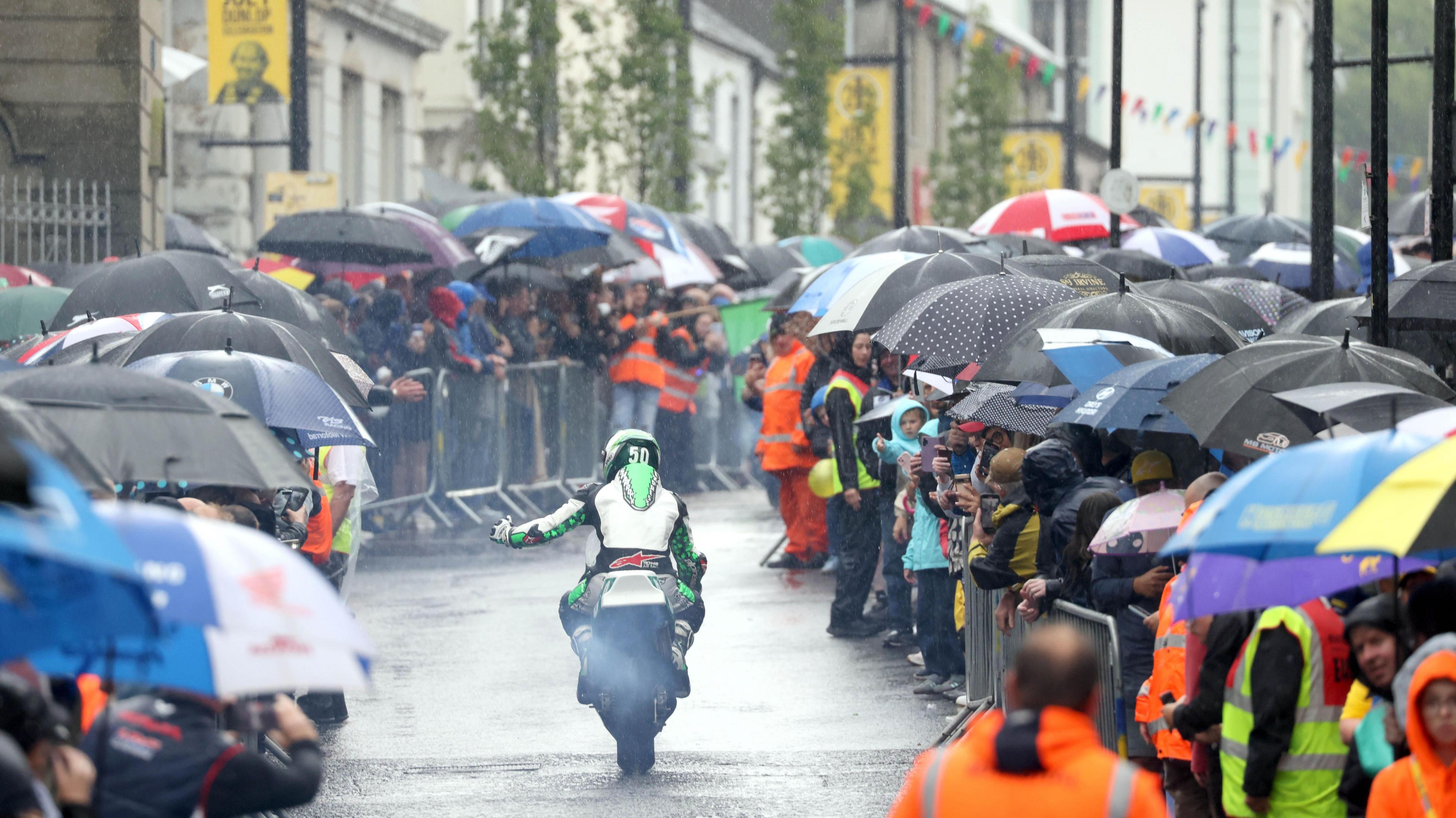 Streets are lined with people holding umbrellas. There are hundreds of people watching a motorcyclist driving through the centre of the road. Rain is bouncing off the road. The riders protective suit is lime green and white.