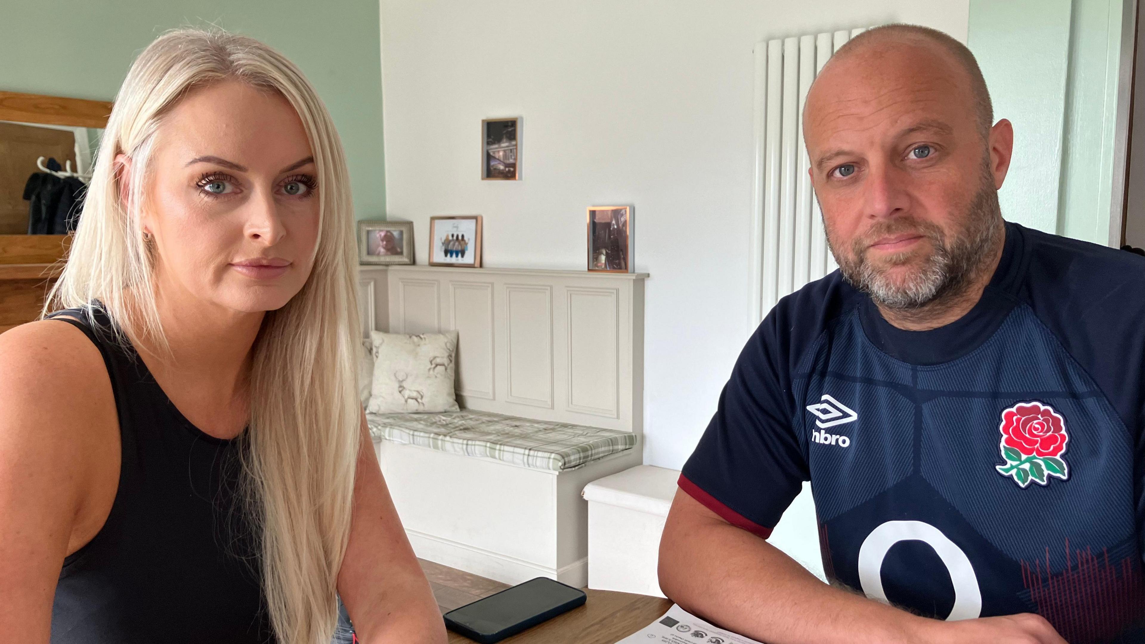 Emma and James Richardson sit at a table in their smartly decorated home. Emma has long blond hair and is wearing a black sleeveless top. James has short fair hair and a neatly trimmed beard and is wearing a blue England rugby shirt. 