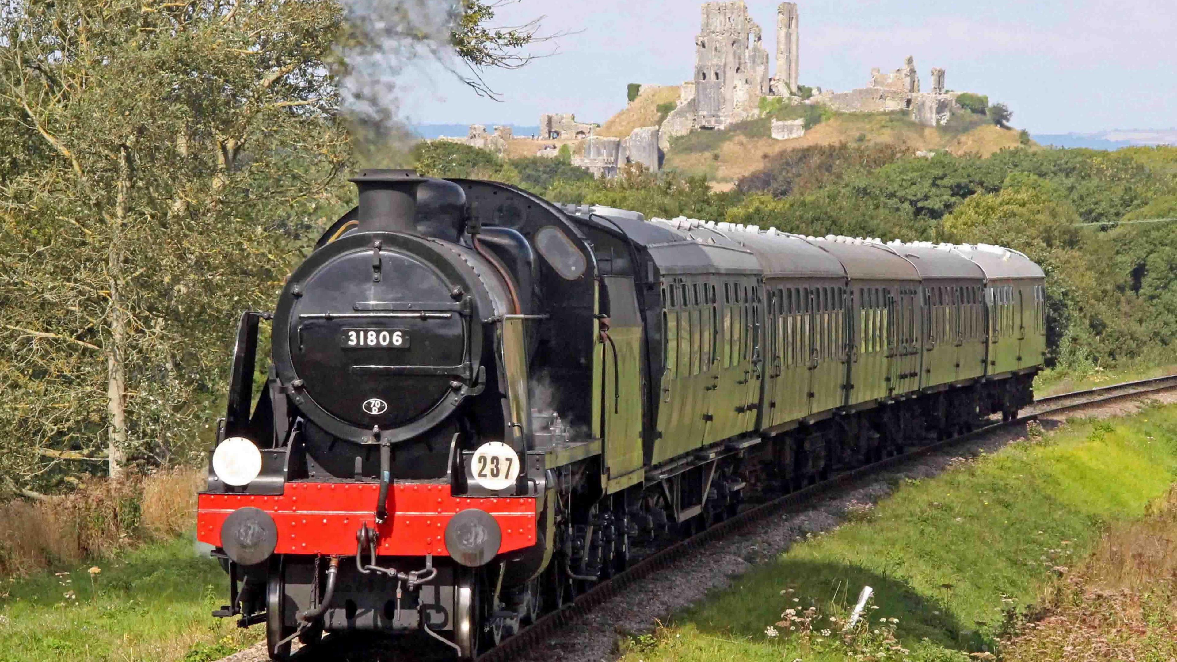 A black steam train moving along a railway track with the ruins of Corfe Castle on a hill behind it.