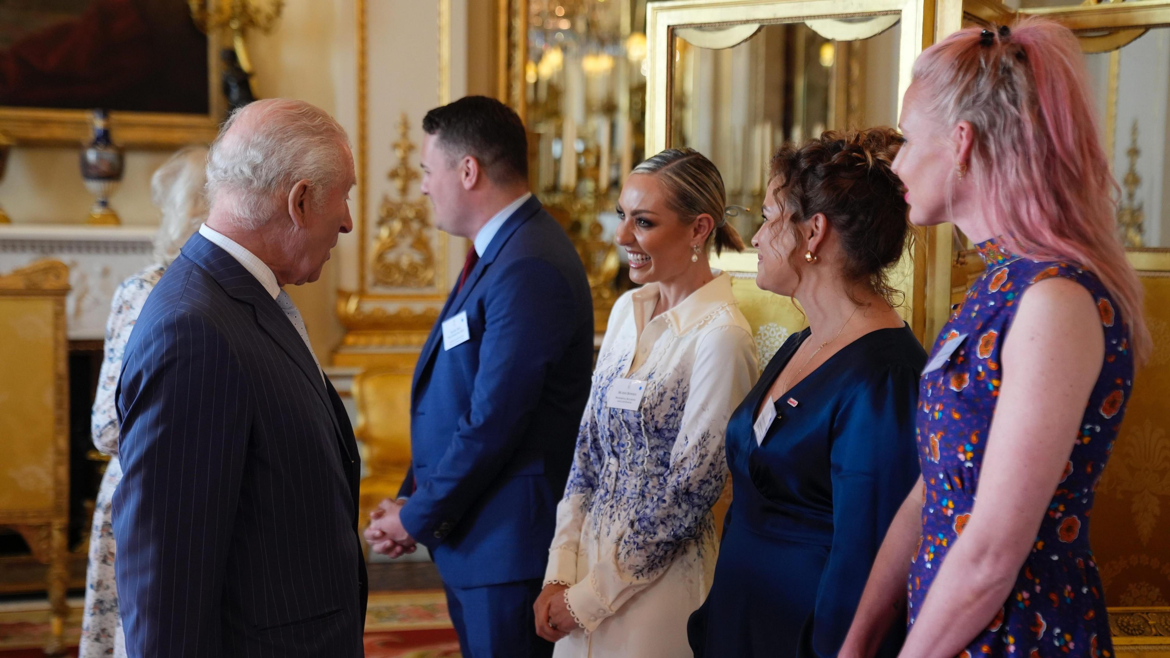 King Charles III looks at Amy Dowden who is wearing a white and blue dress. Behind, furniture is adorned in gold in the halls of Buckingham Palace.