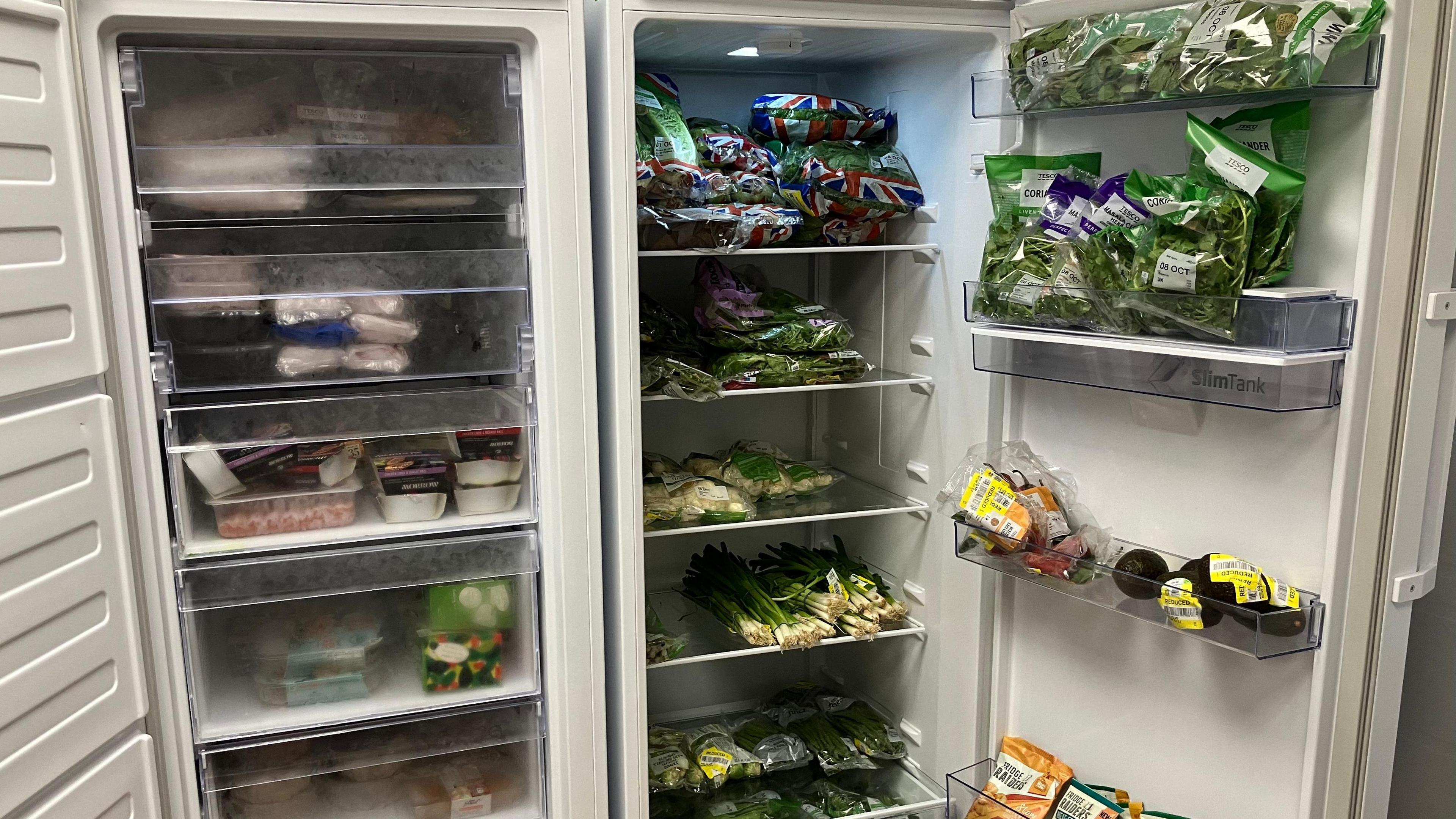 A large free standing fridge fully stocked with vegetables such as spring onions, avacado, brussel sprouts and lettuce.  Its doors are open.  Beside the fridge is a large freezer section full of frozen meat, vegetarian and vegan produce.