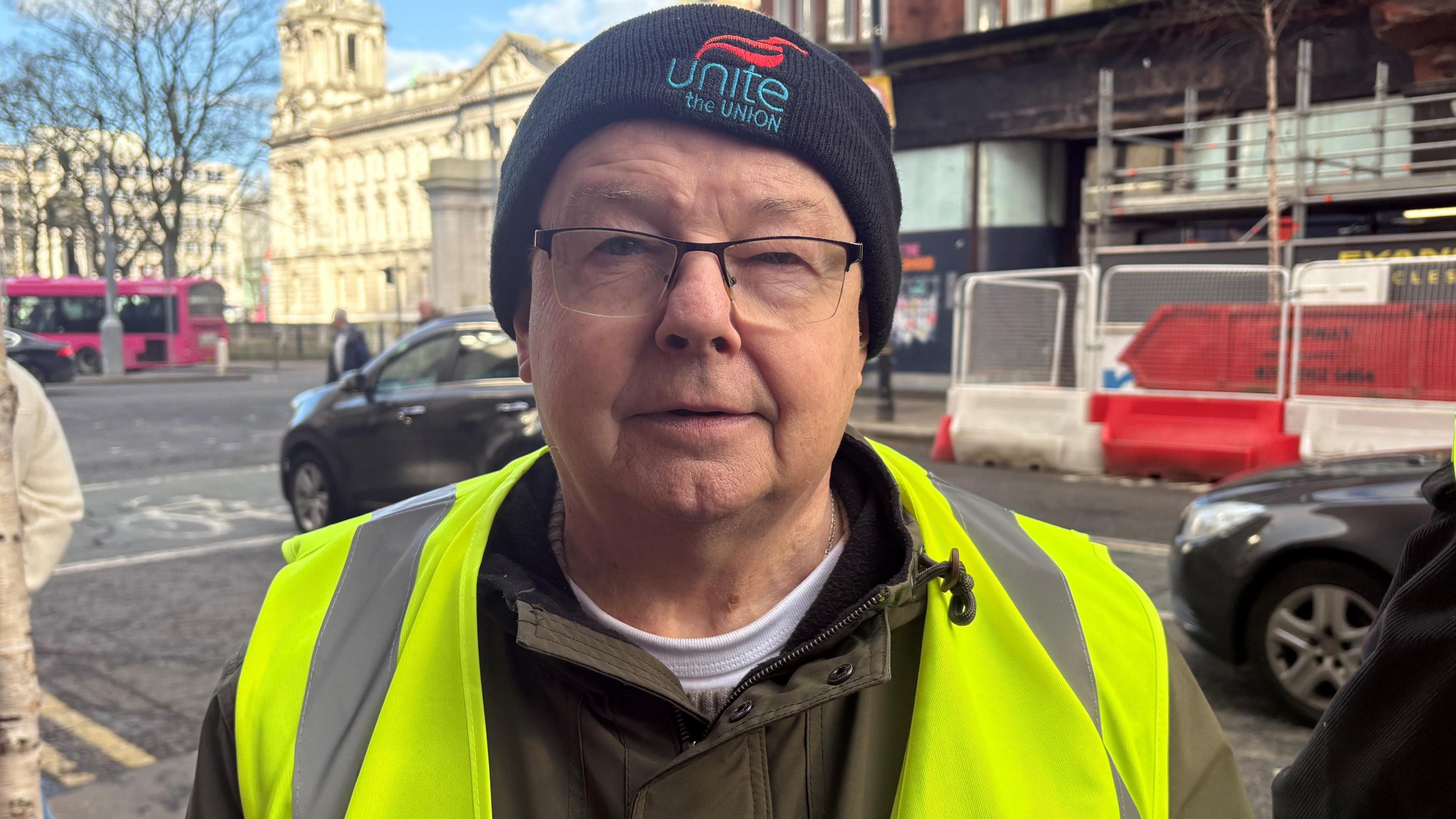 Denis is wearing glasses and a high vis jacket and looking into the camera. He's standing on a street with cars passing him on the road behind him. He's wearing a hat that says Unite the union. 